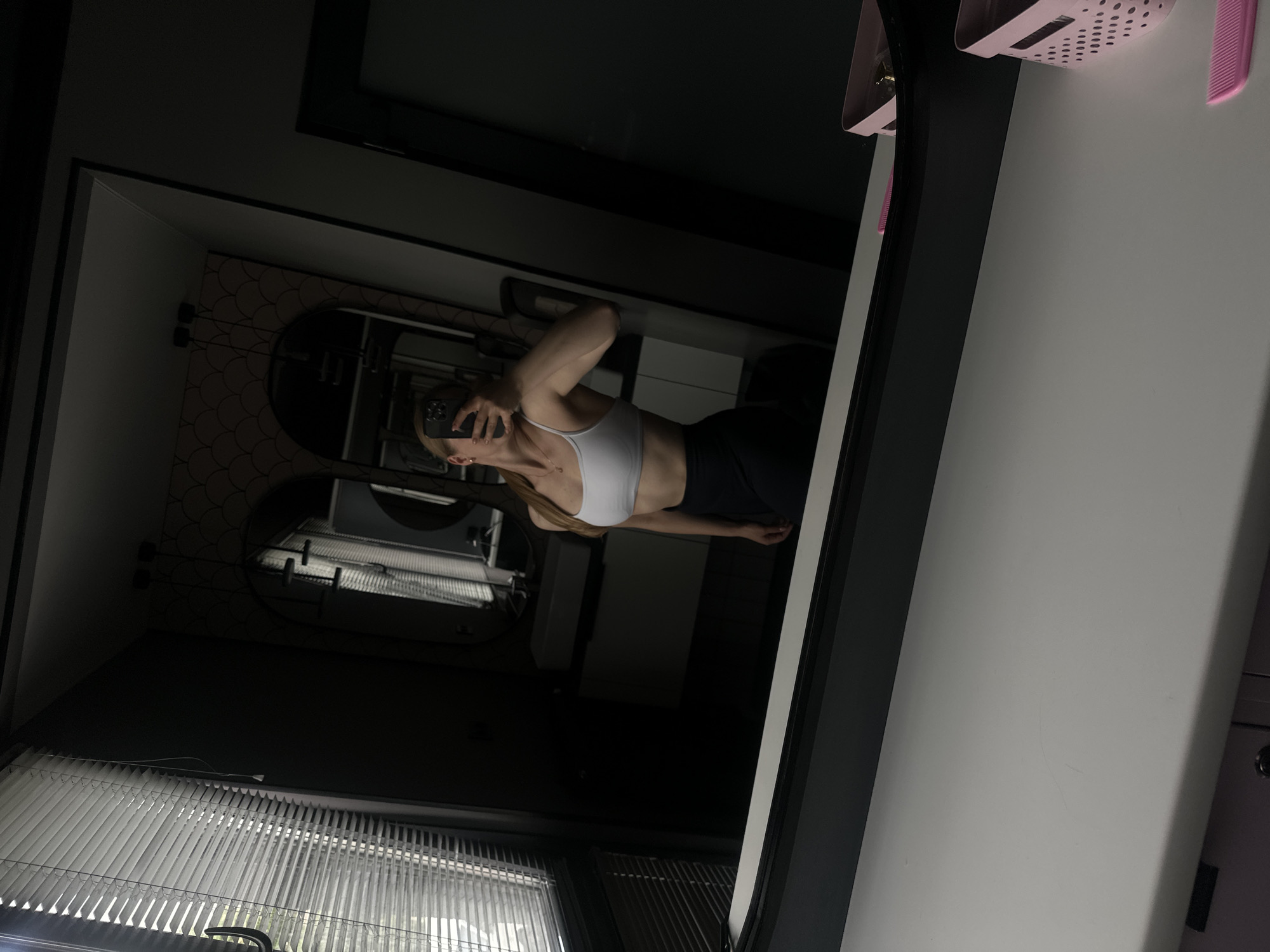 An indoor, dimly lit scene depicts a young adult female taking a mirror selfie within what appears to be a bathroom or dressing area. The woman, with light-colored hair pulled back, wears a white sports bra and dark leggings, holding a dark smartphone to capture her reflection in a large, black-framed mirror. Her face is obscured by the phone. The mirror reflects a portion of the room behind her, revealing a wall with dark, patterned wallpaper, two black pendant light fixtures, and an arched mirror situated above a white rectangular sink and dark cabinet. To the far left, a window with partially open white horizontal blinds allows some ambient light into the room. A white desk or vanity surface in the foreground displays a faint linear mark, a light pink perforated basket containing indistinct items, and a pink comb. On the far right, a dark doorway or frame is visible. The specific geographical location of Ternopil, Ukraine, cannot be ascertained from the visual evidence within the image.
