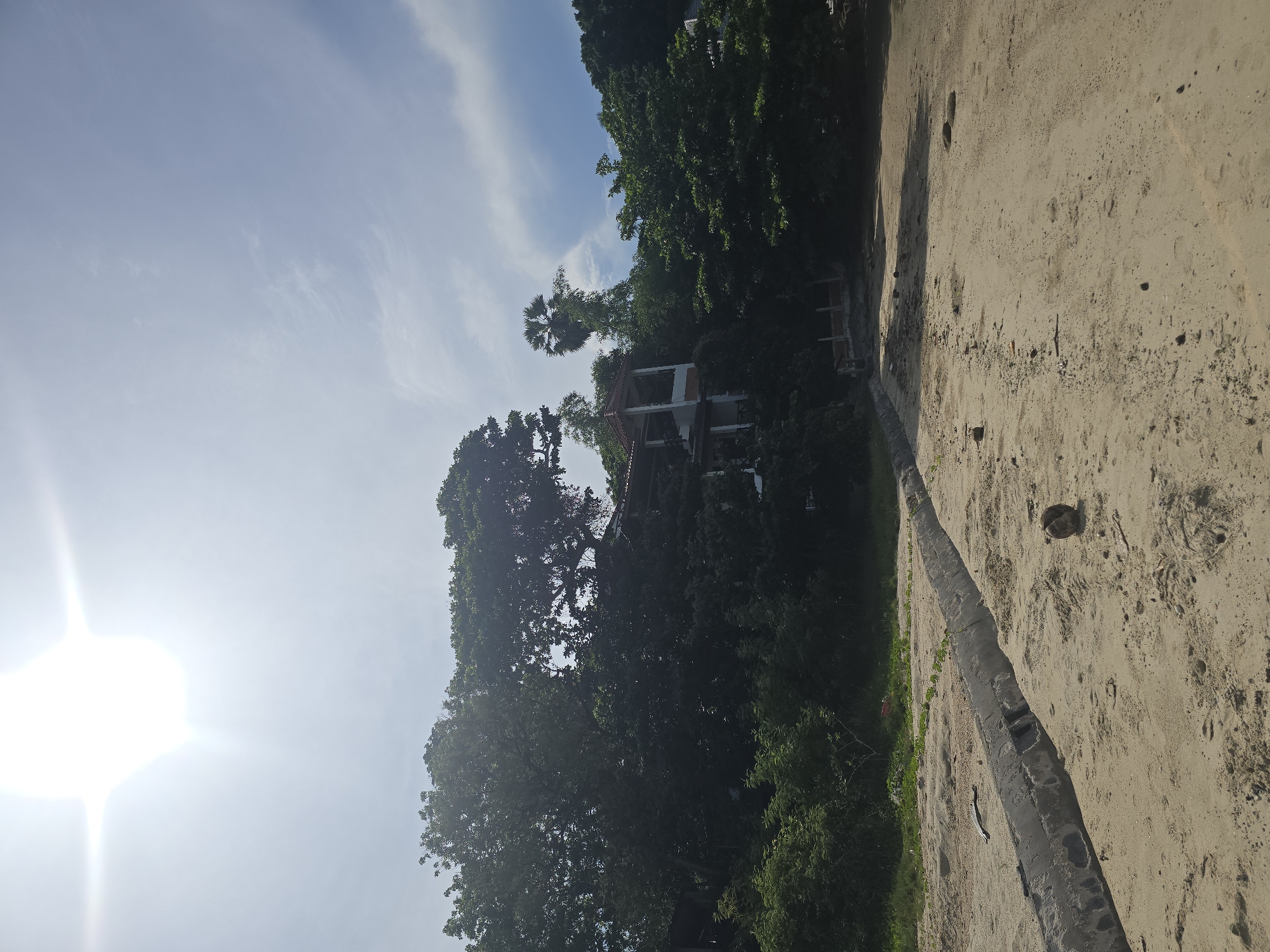 A sandy beach in Baan Chaweng Noi, Thailand, is pictured under a bright, hazy sun and a sky with light clouds. In the foreground, a concrete structure, possibly a drainage channel, runs along the sand. Several coconuts are scattered on the beach, along with some debris and vegetation. Behind the beach, a dense collection of trees and foliage covers a hillside. A building with a white facade and a red-tiled roof is visible amidst the trees, with multiple levels and open balconies. The environment appears to be a coastal area, possibly with a resort or residential development integrated into the natural landscape. The image depicts a serene, sunny day on a tropical beach.