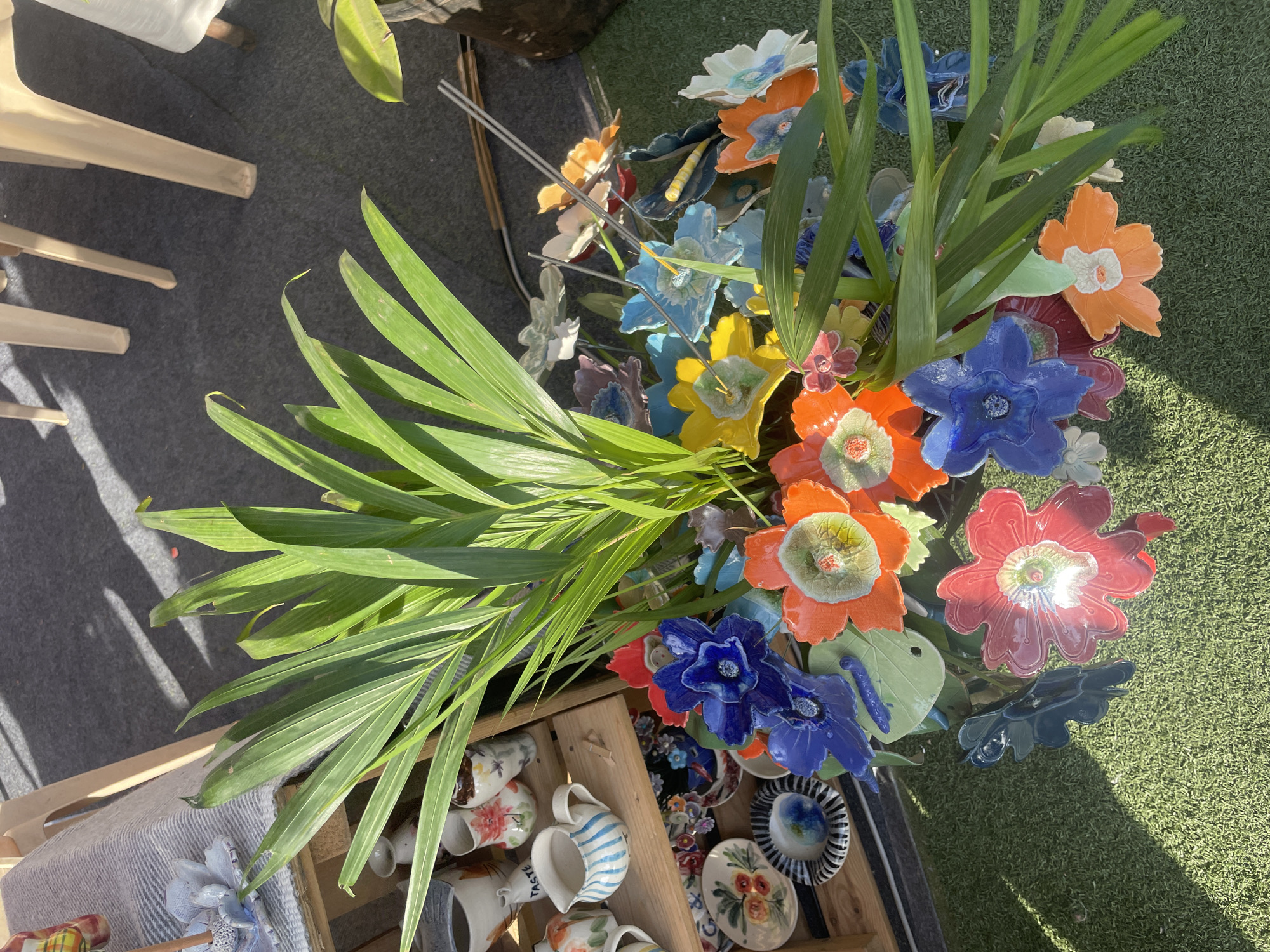 The image captures a vibrant display of ceramic flowers arranged with leafy greenery, likely at an outdoor market or craft fair in Mumbai, India. The arrangement is in a wooden crate alongside ceramic mugs and plates, suggesting a vendor's stall. The ceramic flowers are brightly colored in blue, orange, red, and yellow, with details like petals and centers clearly visible. The leaves are long and green, adding a natural element to the display. The setting appears to be a sunny day, as indicated by the shadows cast on the green artificial grass and grey carpet underneath. The foreground includes glimpses of other items for sale, like more ceramic pieces and the legs of a white plastic chair. Overall, the scene is a commercial setting displaying ceramic crafts.