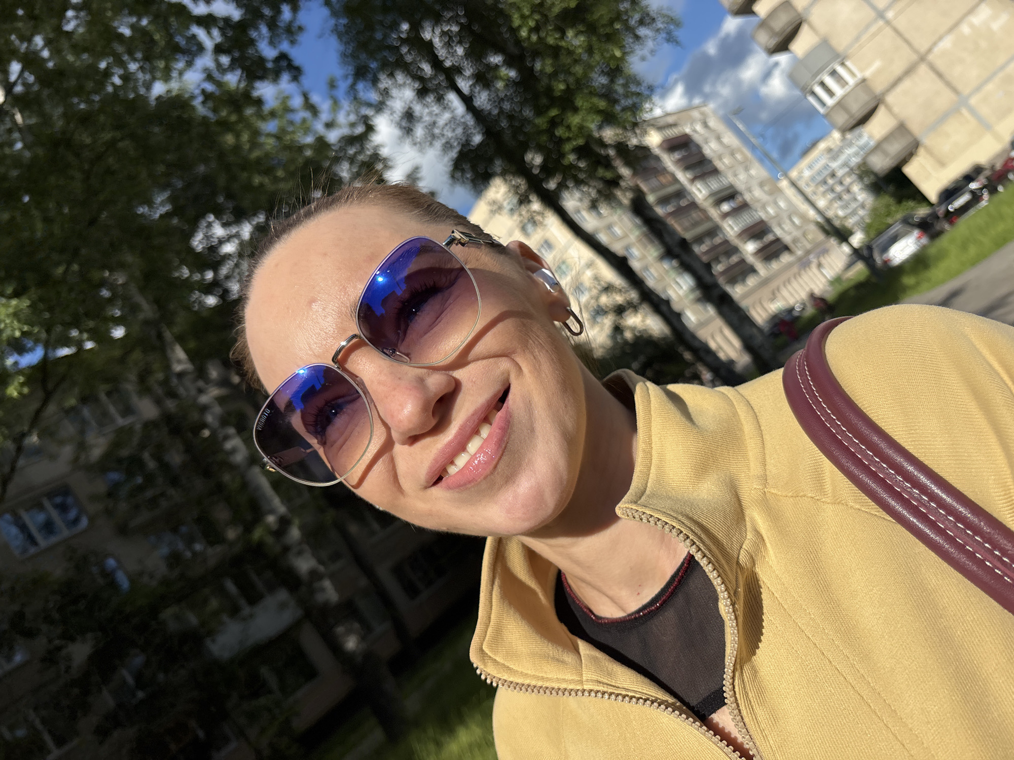 A close-up, head-and-shoulders selfie shows a woman wearing oversized sunglasses with purple lenses and a yellow zip-up jacket. She is smiling, with her mouth slightly open, revealing her teeth. The background is blurred, showing trees, a building, and a cloudy sky. The lighting suggests it is daytime. The location appears to be an urban environment, possibly in Russia, based on the architectural style of the building and the presence of trees commonly found in Russian cities.