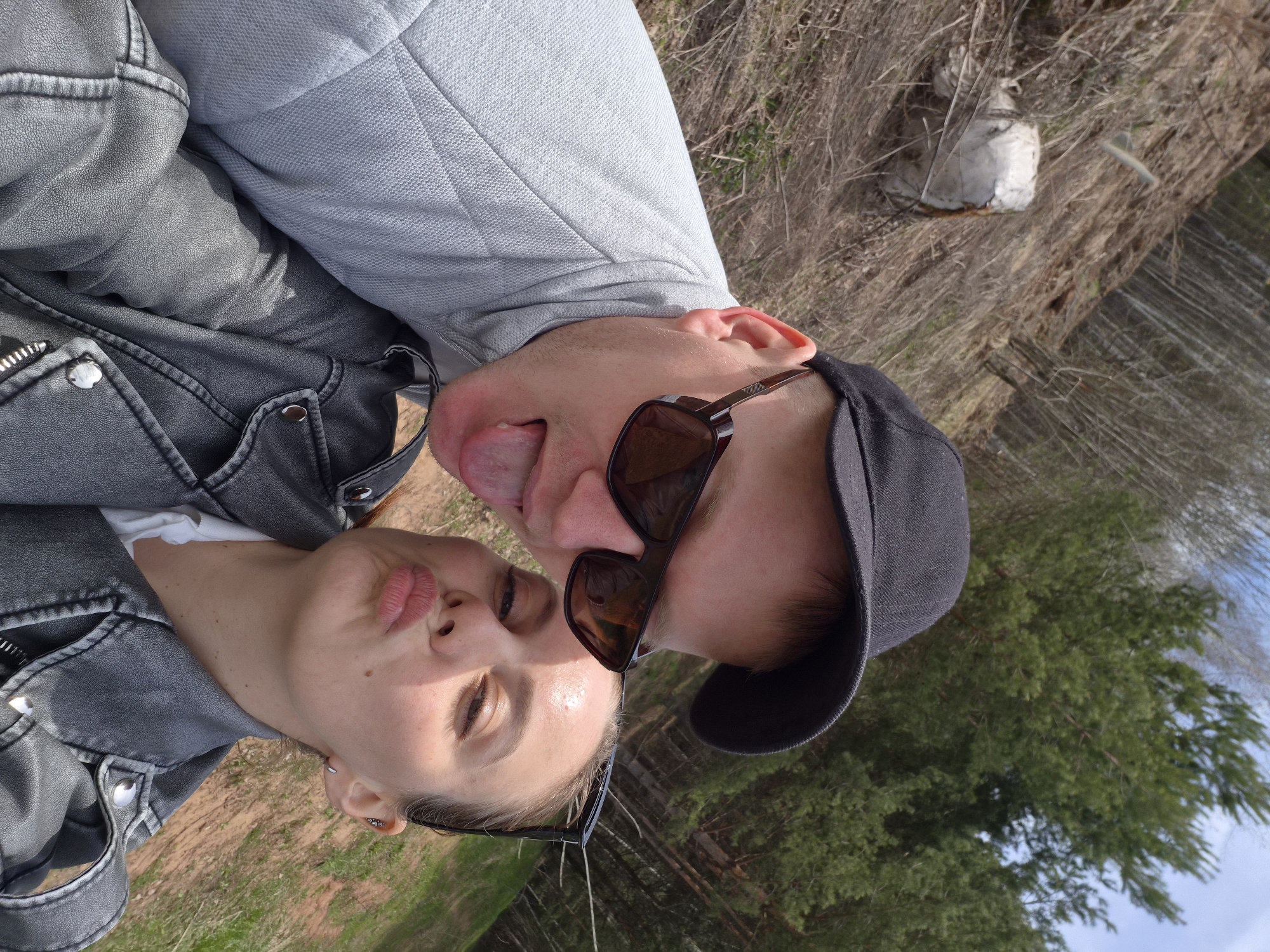 A playful, close-up selfie captures a man and a woman in an outdoor setting. The image, oriented in portrait mode, shows the woman on the left and the man on the right, with their faces close together.
The woman has light skin and is dressed in a dark grey jacket, possibly faux leather, worn over a white top. Her light brown hair is pulled back, and a pair of sunglasses rests on her head. Her lips are puckered in a kissing gesture, and her eyes are mostly closed. A small silver earring is visible in her left ear.
To her right, the man wears a light grey long-sleeved shirt and a black baseball cap. His eyes are obscured by dark sunglasses. He is playfully sticking out his tongue. His left ear is visible, and he appears to be clean-shaven.
The background is a blurred natural landscape. Behind the woman on the left, dry, reddish-brown earth and some sparse green grass are visible. Behind the man on the right, tall green pine trees extend upwards against a light blue sky. In the upper right area of the background, there is an expanse of dry brown brush, and a crumpled white object, possibly a plastic bag or trash, is visible.
The bright, direct sunlight indicates it is daytime with clear weather. The overall mood conveyed by their expressions is lighthearted and affectionate. The setting suggests a natural or rural area, consistent with the Chaikovsky, Russia context. There is no visible text in the image.