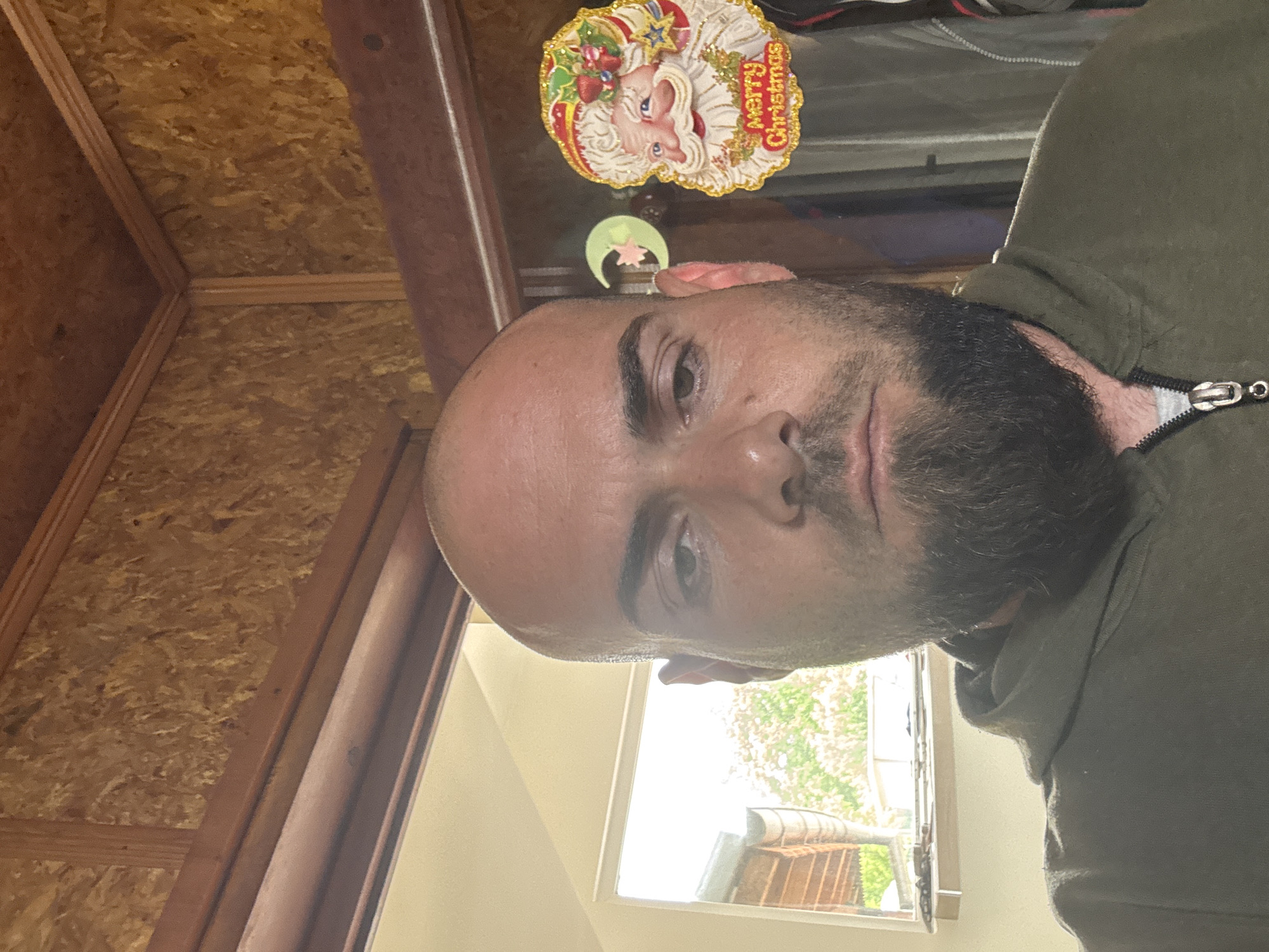 A medium shot of a bald, bearded man wearing a green hooded sweatshirt. The image appears to be a selfie taken inside what may be a shed or storage space with unfinished wood walls. A Christmas decoration hangs near the top of the frame. A window provides light to the right of the man, showing trees and a portion of a building outside. Sarnevo, Bulgaria cannot be confirmed.