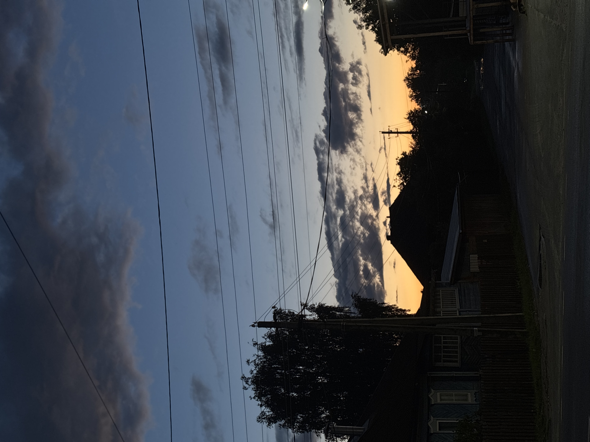 This media file captures a tranquil street scene at either dusk or dawn, featuring a striking gradient sky. The sky transitions from a deep, dusky blue on the left to a vibrant orange and yellow hue towards the right horizon. Scattered across the sky are various clouds, some dark and heavy, others lighter and wispy, creating a dynamic visual texture.
Several parallel power lines stretch horizontally across the frame, cutting across the sky. Silhouetted against the colorful sky are dense clusters of trees, predominantly on the right side and surrounding a utility pole in the middle-right. Another utility pole with a visible light source (likely a streetlamp) is present in the upper right.
In the lower right part of the image, dark silhouettes of buildings and structures are visible, lining what appears to be a road or paved area. One identifiable structure is a traditional wooden house, featuring distinct, ornate window frames. No people or specific activities are depicted. The scene conveys a peaceful, atmospheric moment, likely in Krasnye Baki, Russia, given the location context. No visible text is present.