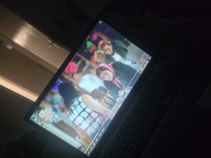 The image displays a laptop screen actively playing a video in a dimly lit indoor environment. No specific features identifying a location within Kaiserslautern, Germany, are discernible.
**Subjects/Objects:**
The primary subject is a laptop computer, with its screen prominently featured, showing a scene from a video. The laptop's keyboard is visible in the lower foreground. The video content includes multiple individuals. A person with long, dark hair wearing a white and black checkered or plaid top is seen from the back or side, facing away from the viewer. To the lower right of this person, a woman wearing a pink head covering (possibly a scarf or hijab) is visible, looking towards the right side of the screen. Other blurred figures and colorful decorations, resembling pompoms or balloons, are present in the background of the video. A 'B' logo is visible in the upper left corner of the video playback. A playback progress bar at the bottom of the screen indicates a duration of "03:15 / 07:02", alongside partially visible playback controls (play/pause, volume, settings, full screen icons). In the physical background of the room, a light-colored door with a dark handle is visible to the upper right of the laptop screen. A dark, possibly textured surface, consistent with bedding or fabric, is visible to the right of the laptop.
**Environment:**
The overall scene is dimly lit, suggesting low ambient light in the room. The primary light source appears to be the laptop screen itself. The door in the background suggests an interior residential or office space.
**Actions/Interactions:**
The laptop is engaged in playing video content. The individuals depicted in the video appear to be within a social setting, possibly interacting or observing.