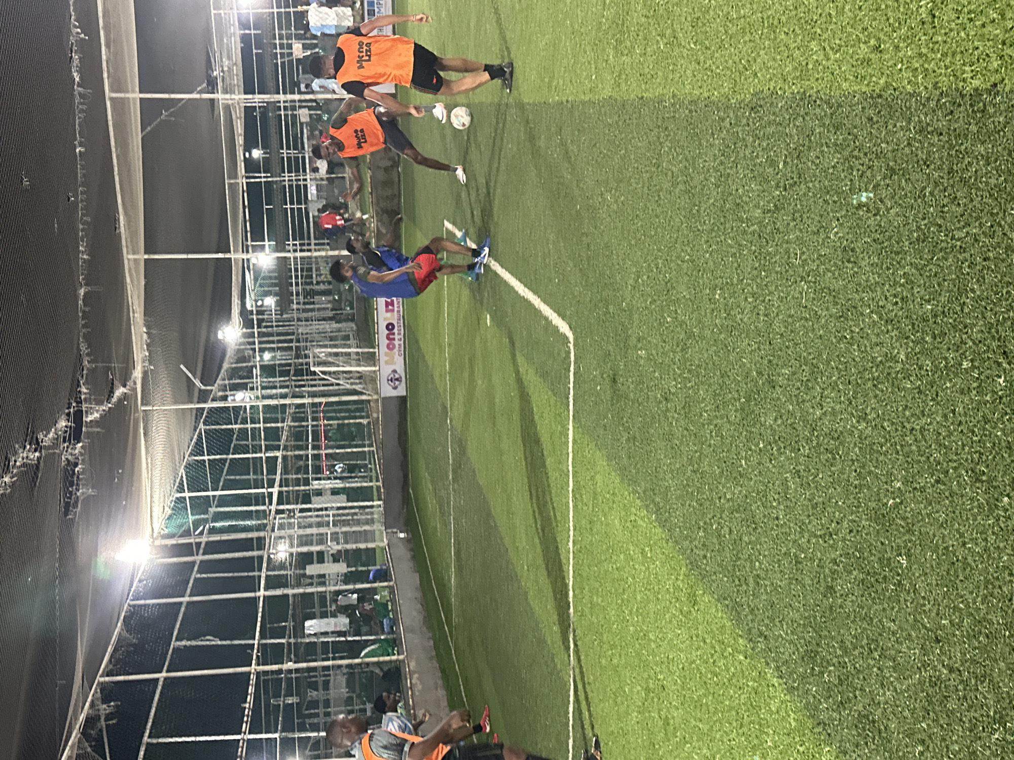 This image depicts a football match at an indoor artificial turf pitch in Abuja, Nigeria, likely during the evening or night, as evidenced by the artificial lighting. The pitch has white boundary lines and a net enclosure surrounds it. In the foreground, a person in an orange bib with "Mono Liza" printed on it is on the right side of the frame, facing away from the camera and towards the action. To their left, another individual in a similar orange bib is engaged in the game, with their back to the camera, leaning forward and looking at a football at their feet. Further into the pitch, a person in a blue jersey and red shorts is actively playing, looking down at the ball. Another person in a red shirt is visible in the background. Other individuals are casually observing the game from the sidelines. The overall scene suggests a recreational football game taking place at a sports facility in Abuja.