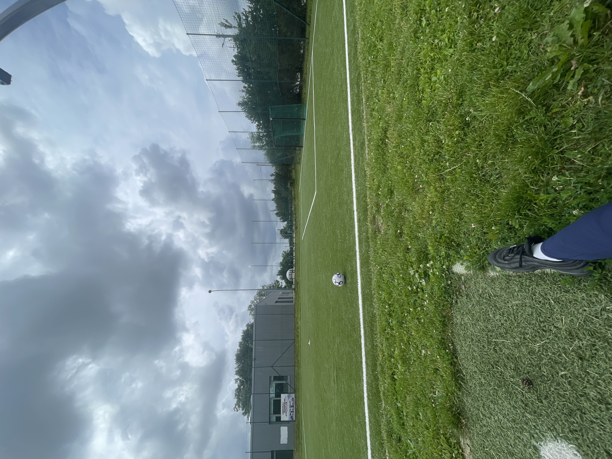 An outdoor shot depicts a soccer field with a low-angle perspective, looking across the green turf. In the foreground, a person's left leg, clad in dark blue pants and a black athletic shoe, is visible. The field is marked with white lines. A soccer ball rests on the grass near the center line. In the background, a grey building with a large sign displaying images of trucks is situated to the left. Beyond the building, a chain-link fence encloses the field, with trees and foliage visible behind it. The sky is overcast with dramatic, dark clouds. A lamppost stands tall near the building, and a soccer goal with a green net is visible on the right side of the field. The overall scene suggests a cloudy day at a sports facility. The location is identified as Jakai, Lithuania.