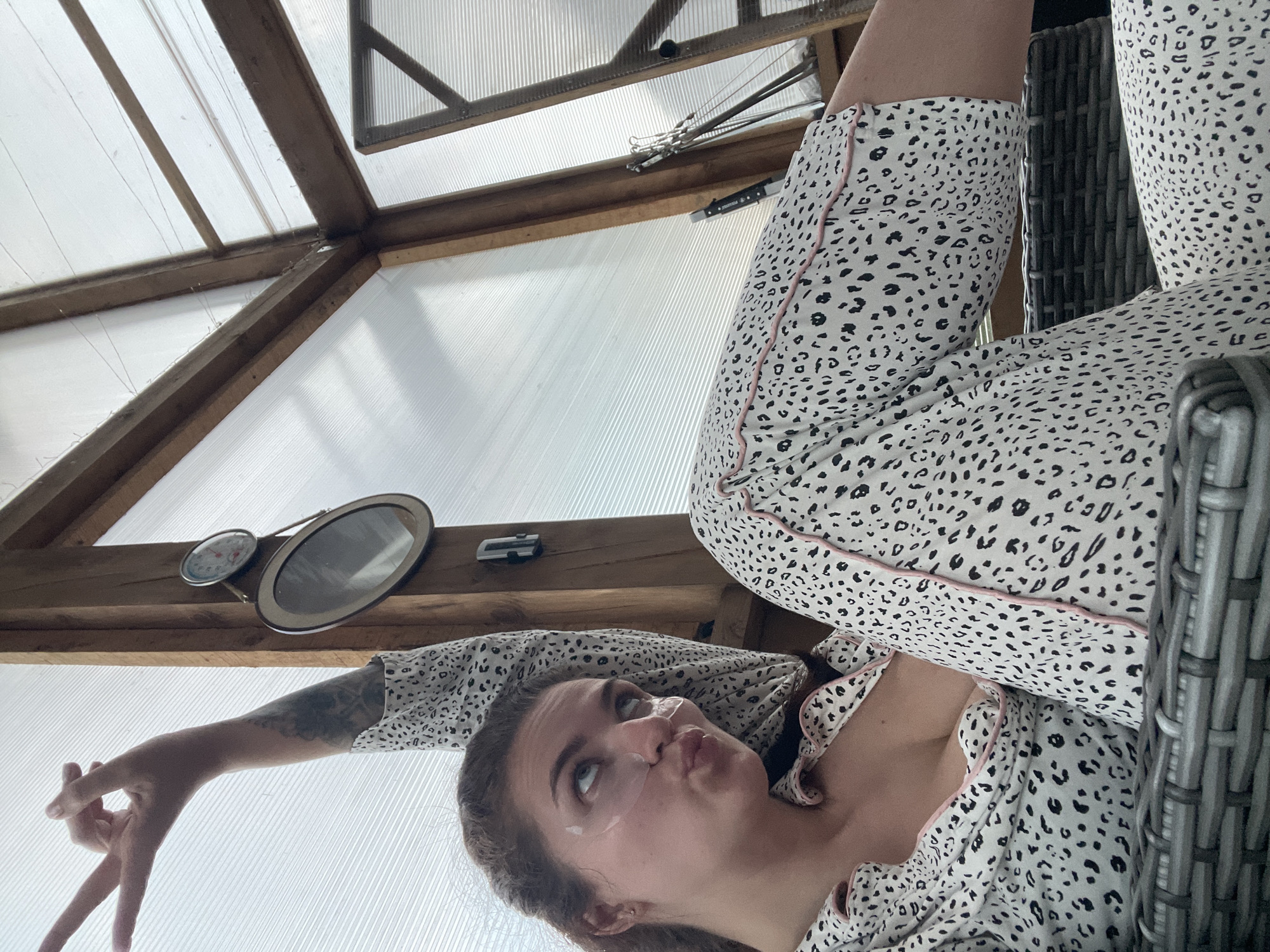 A person wearing spotted pajamas is reclining in a chair in a room with a translucent roof. The person has what appear to be facial patches applied under their eyes and their face is tilted upwards. The person's left arm is outstretched, fingers splayed. A round thermometer and a framed picture are mounted on a wooden beam above the person. The exact location in the city of Мосметаллоконструкция Онт, Russia cannot be determined from the image.