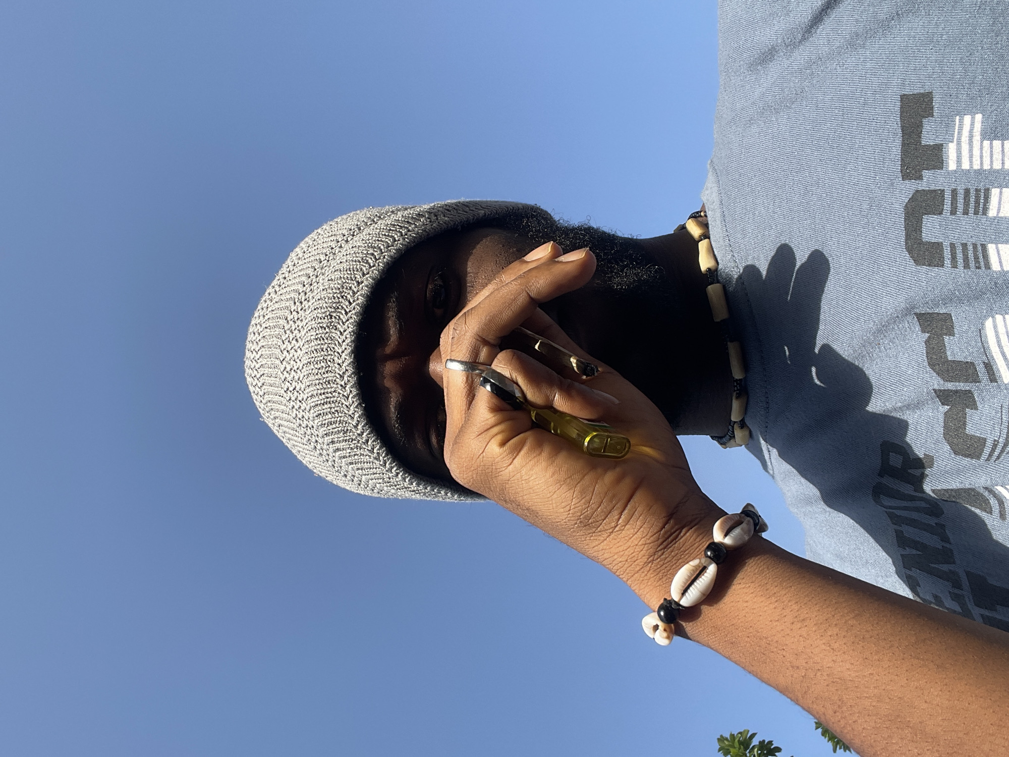 A person stands outdoors in Jos, Nigeria, against a clear blue sky. The person is wearing a grey knitted beanie and a blue t-shirt with black and white graphics. They have a beaded necklace and a bracelet made of shells and black beads on their wrist. Their hand is raised, holding a yellow and transparent object. They also have a ring on their finger.