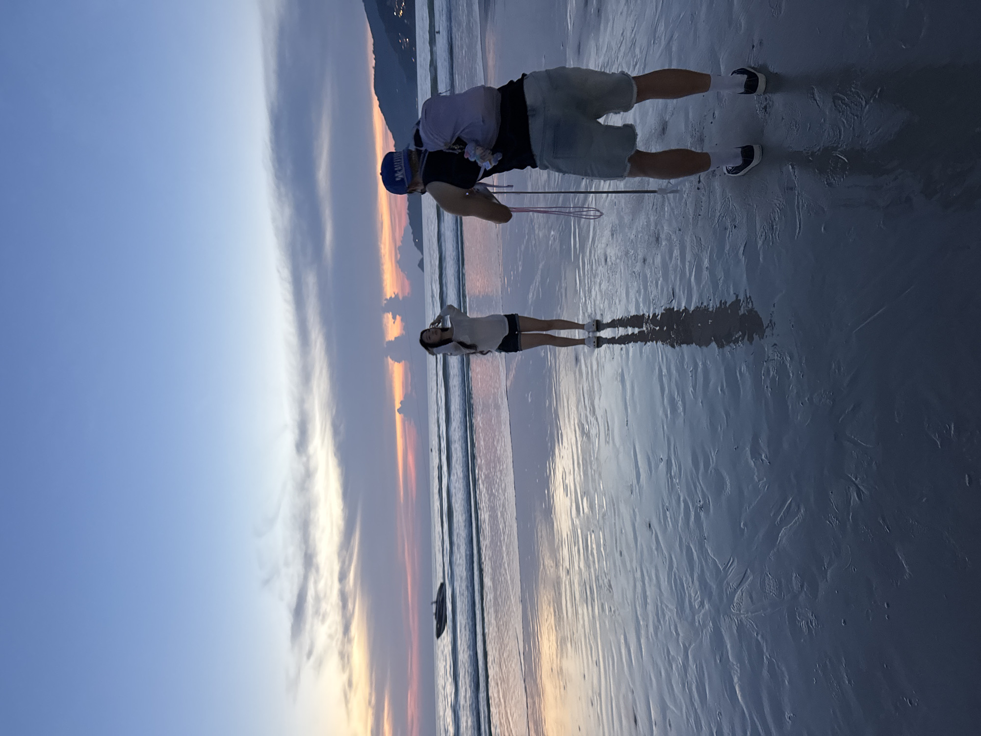 A low-angle, full-shot photograph captures a man and a woman on a beach at dusk, likely in Patong, Thailand. The sky displays a gradient of colors, transitioning from a pale blue at the top to hues of pink, orange, and purple near the horizon, indicative of a sunset. Wispy clouds are scattered across the sky. The wet sand on the beach reflects the sky's colors and the figures.
In the foreground, a man stands facing away from the camera, wearing a black tank top, light blue denim shorts, white socks, and black sneakers. He is wearing a blue baseball cap and has a light purple backpack slung over his right shoulder. He holds a pink pole with a thin string attached to it, possibly for photography or videography. His reflection is visible on the wet sand.
Further back, a woman stands facing the man, her hands near her head, appearing to be posing for a photograph. She is wearing a light-colored, long-sleeved top and dark shorts. Her reflection is also visible on the wet sand.
In the middle ground, a speedboat is visible on the ocean, creating small waves. The water's surface is choppy. In the far background, hills covered in vegetation and dotted with lights are visible, suggesting a coastal town or city. The overall scene is serene, with the fading light creating a tranquil atmosphere.