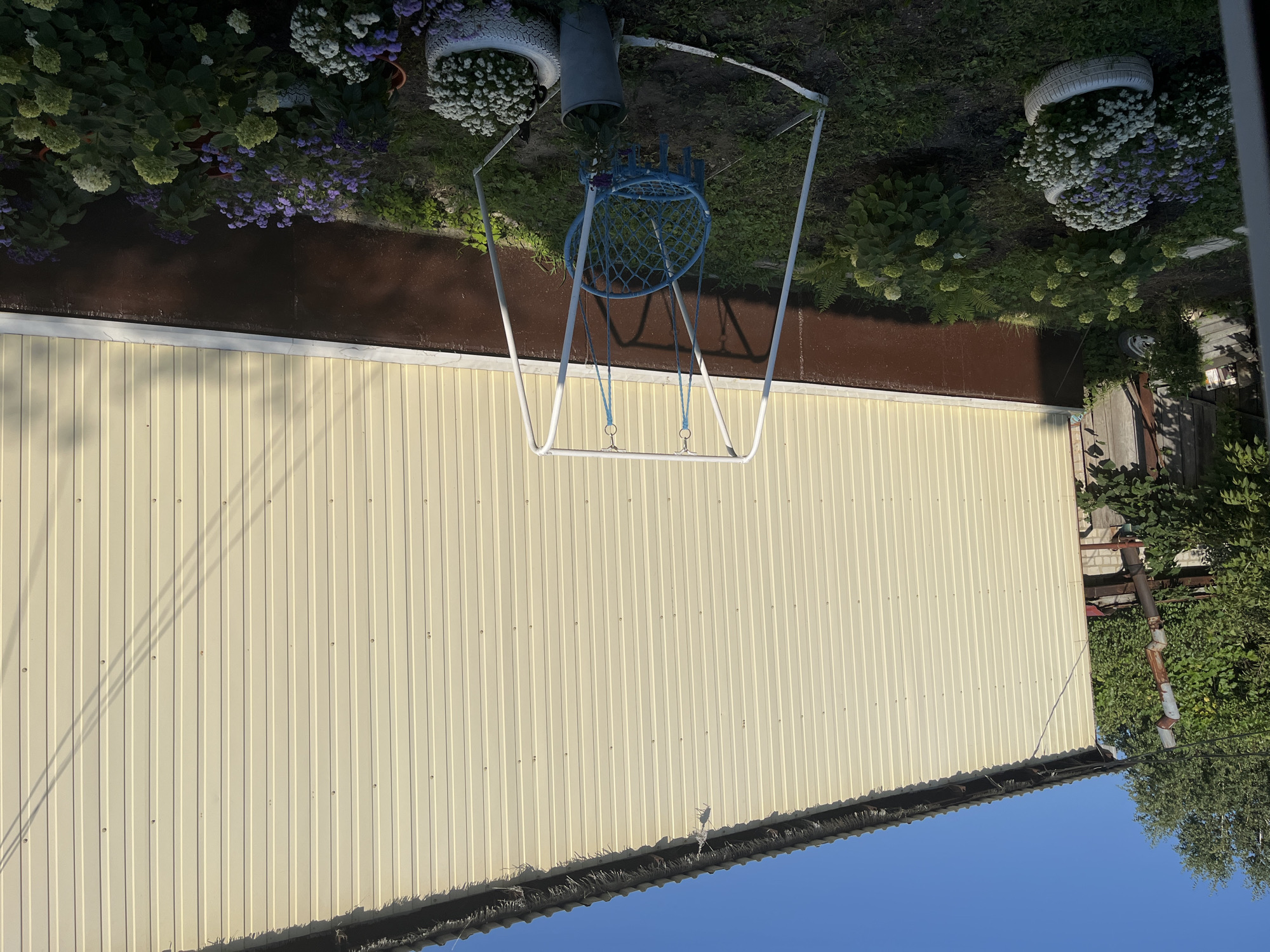 The image depicts a backyard scene in Saratov, Russia. A light-colored corrugated metal building forms the backdrop. In front of it, a white metal swing set with a blue woven seat hangs, situated in a garden area. The garden contains various flowering plants with white and purple blooms, some planted in repurposed tires. Green foliage and trees are visible to the left of the building, and a rusted downspout is attached to the structure. The sky is clear and blue. The scene is illuminated by natural sunlight, casting shadows on the building.