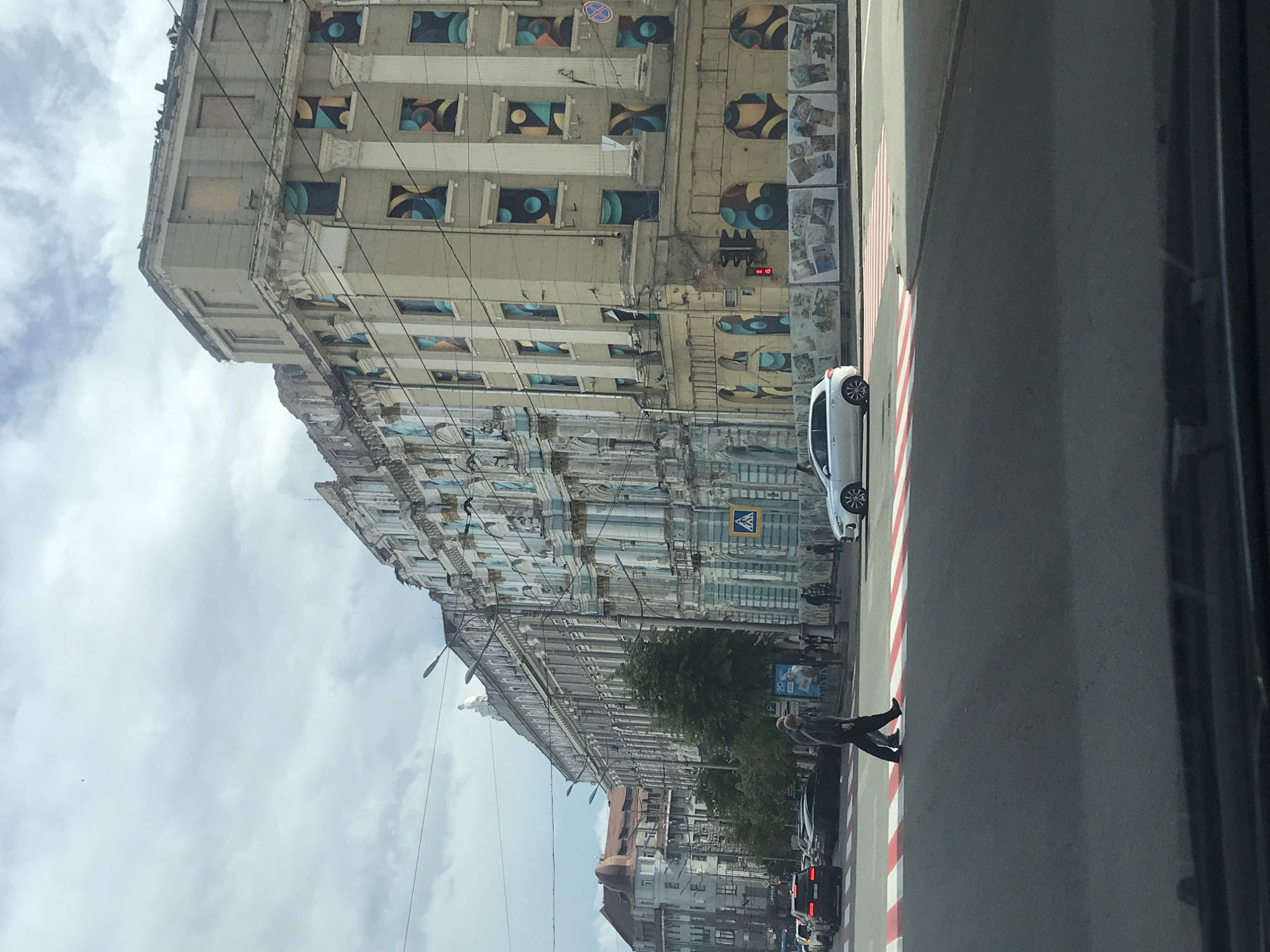 The image shows a street scene in Kharkiv, Ukraine, dominated by a large, ornate building with a painted façade. The building has multiple stories, and the windows on the upper floors are decorated with geometric patterns. The lower floors are heavily ornamented with classical architectural details. A white car is parked along the curb, and people are walking on the sidewalk and crossing the street. A pedestrian crosswalk is marked with red and white stripes. The sky is overcast with clouds. The image appears to have been taken from inside a vehicle, as part of the car's interior is visible on the right. Other buildings are visible further down the street.