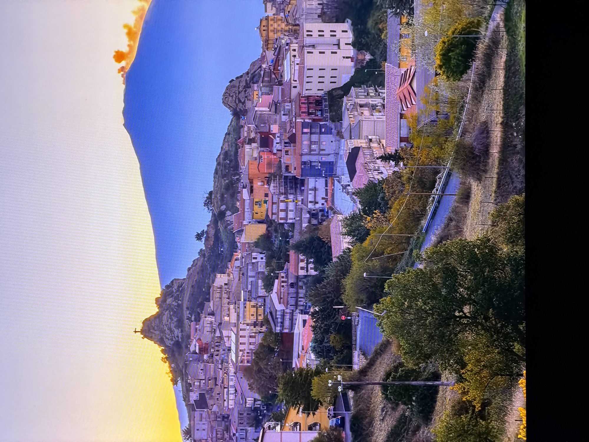 The image displays a panoramic view of a hilltop town nestled against a large mountain at sunset. The town features a dense cluster of buildings with diverse architectural styles and pastel-colored facades, built into the side of a steep incline. At the summit of a rocky outcrop above the town, a silhouette of a cross is visible against the bright sky. The mountain behind the town is a deep blue, with a plume of orange and red smoke erupting from its peak, suggesting volcanic activity. The foreground shows a winding road with sparse vegetation and trees, some of which display autumnal colors. The sky is a gradient of pale yellow and white near the horizon, transitioning to a clear blue higher up. There are no people or vehicles clearly visible in the scene, making it a static landscape. This description is unrelated to Chirchik, Uzbekistan.