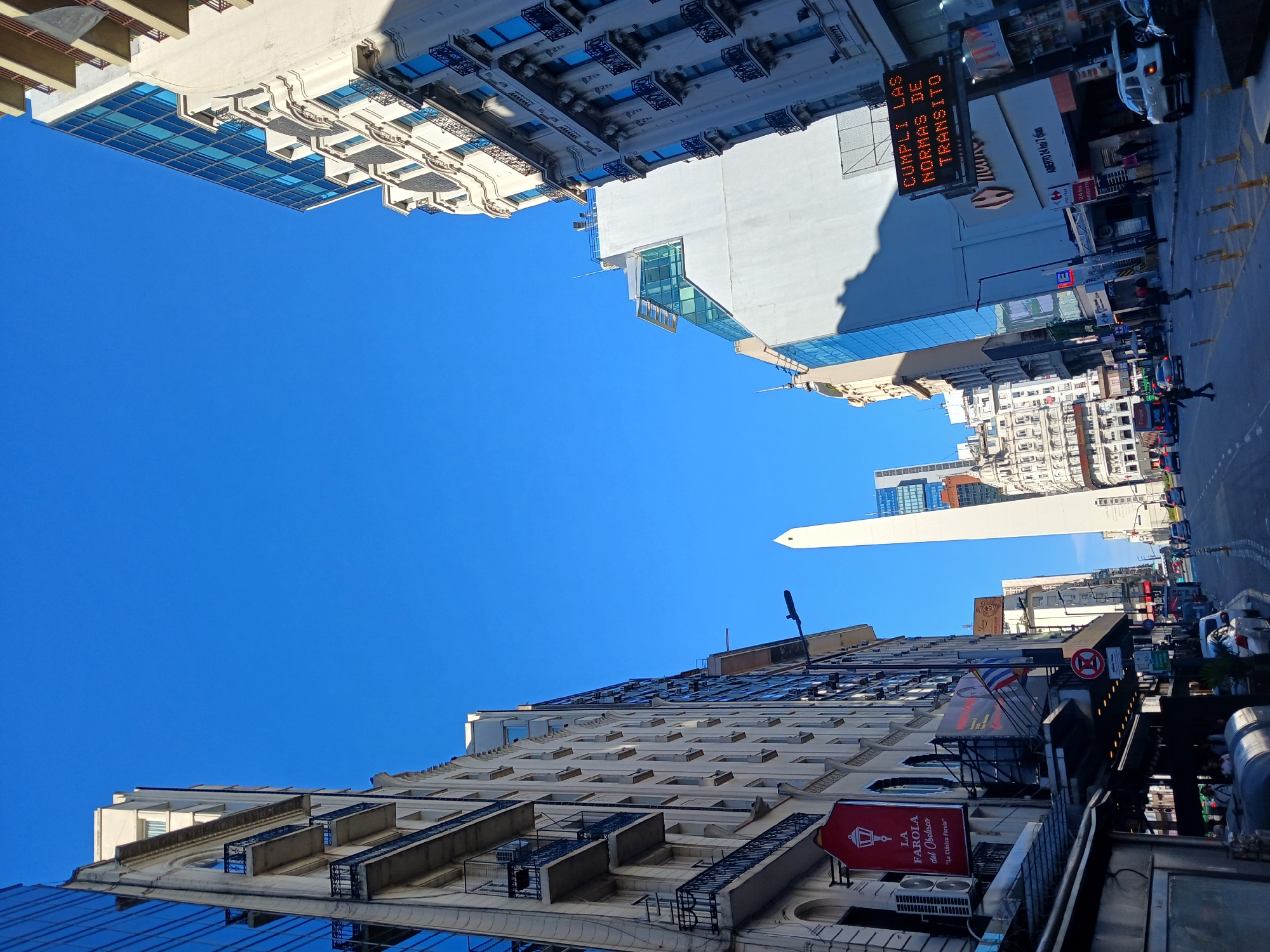 The image captures a view of the Obelisco de Buenos Aires in Argentina, framed by the surrounding buildings and a clear blue sky. The perspective is tilted, looking upward towards the monument. In the foreground, buildings line the street, some displaying signage and balconies. The street below shows traffic and pedestrians. A prominent digital sign displays the message "CUMPLI LAS NORMAS DE TRANSITO" in Spanish, meaning "Comply with traffic regulations". A red sign for "LA FAROLA del Obelisco" is visible on one of the buildings.