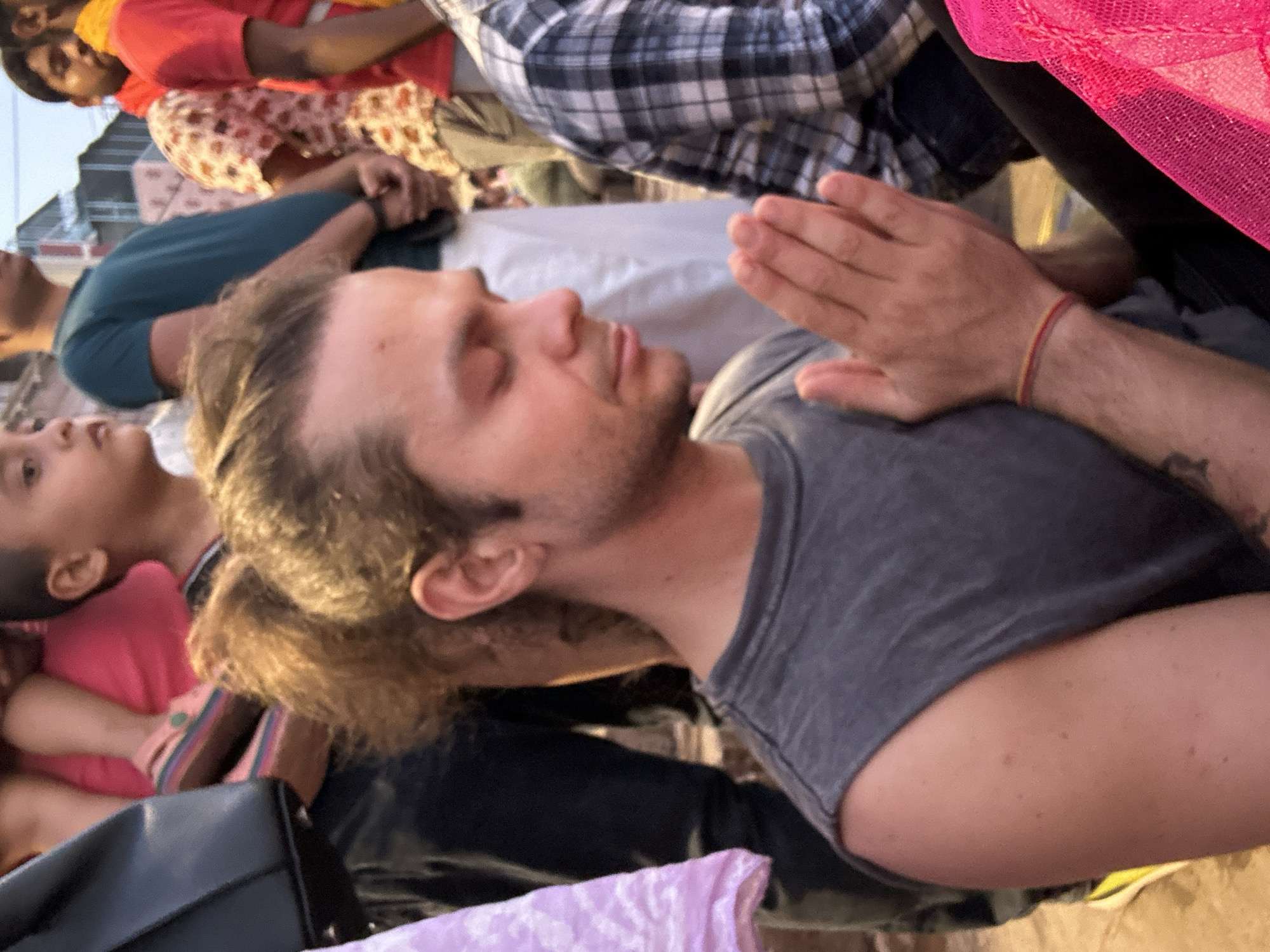 The image captures a medium shot of a man with fair skin and light brown hair, styled in a low ponytail, in a meditative posture with his eyes closed and hands pressed together in prayer. He is wearing a grey t-shirt and a red and yellow bracelet on his wrist. A tattoo is visible on his arm. The photograph appears to be taken from a low angle.
In the background, a group of people are visible, including a child wearing a pink shirt and a man in a teal t-shirt. Other people are wearing brightly colored clothing. A black leather bag is also visible. Given the context of the image, it is likely taken in Varanasi, India. The scene suggests a spiritual setting where people are engaged in prayer or meditation.