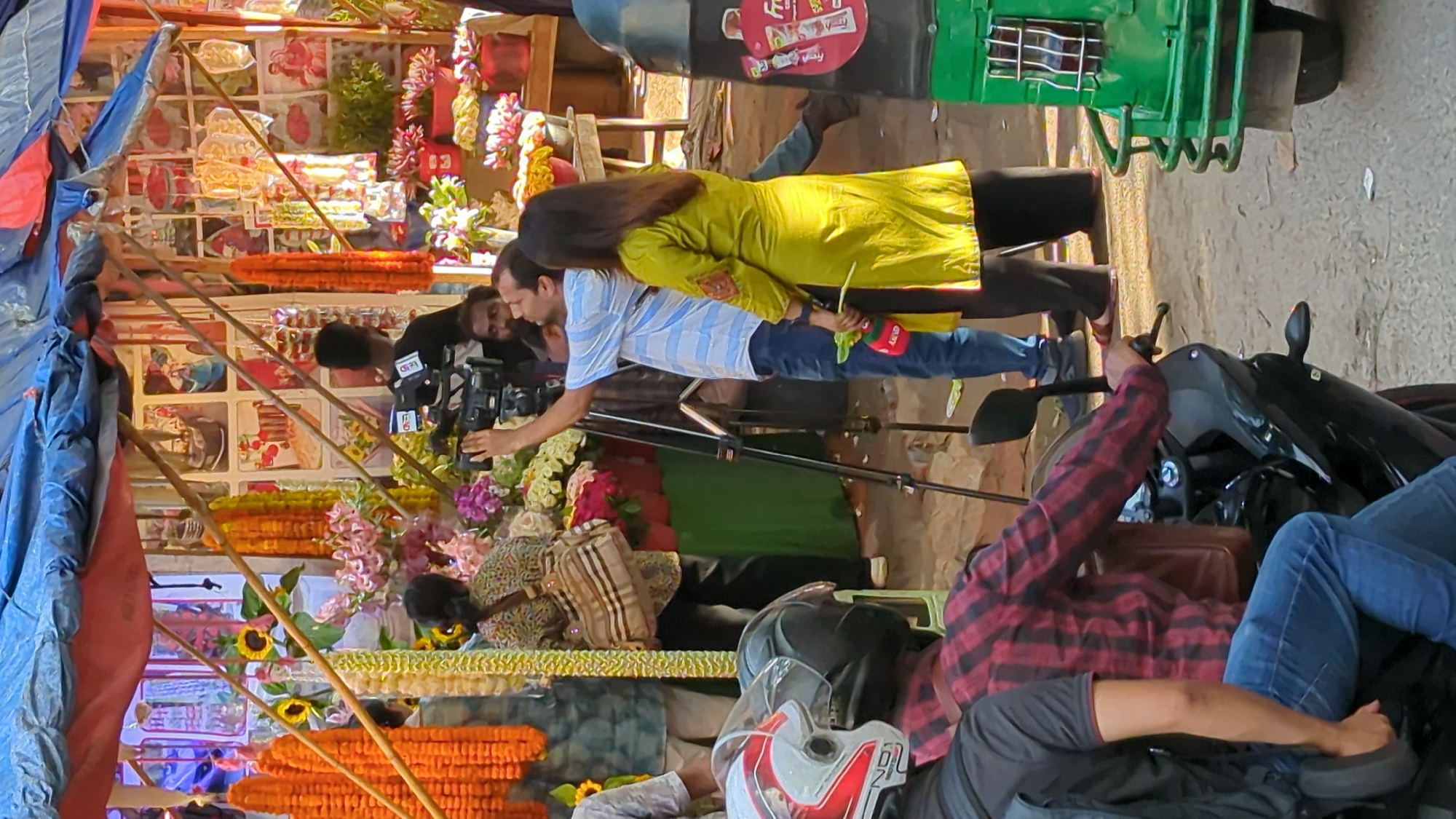 In an outdoor market scene, possibly in Dhaka, Bangladesh, a person is operating a professional video camera mounted on a tripod. The camera operator, dressed in a blue and white striped shirt and blue jeans, is looking upwards. To their right, a person wearing a bright yellow tunic and dark pants is holding a green leafy item and also looking away from the camera. Behind them, a person in a patterned shirt and a large bag slung over their shoulder stands near a display of flowers and garlands. Further back, several other individuals are visible, some tending to the market stalls which are decorated with colorful garlands and various items. In the foreground, two people on a motorcycle, one wearing a red and black plaid shirt, are stopped, suggesting traffic or a pause in movement. A green auto-rickshaw is parked to the right of the frame. The overall atmosphere appears to be that of a busy street market.