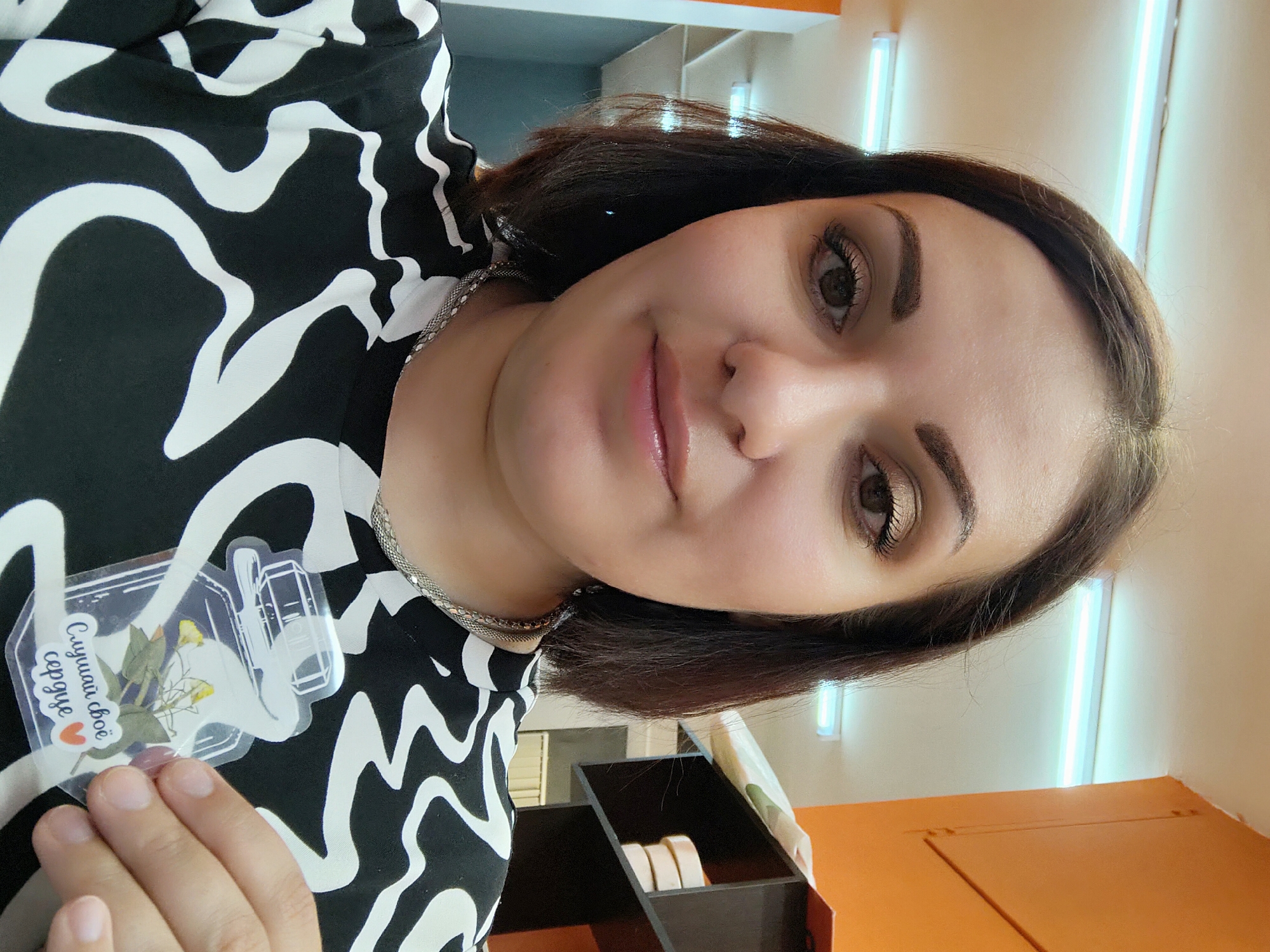 A woman is pictured indoors. She is facing the camera at an angle, with her head tilted slightly to the right. She has short, dark brown hair, light skin, and is wearing neutral makeup. She's wearing a black and white patterned top and a silver chain necklace. A clear sticker is visible on her top, featuring an image of flowers and a Russian inscription with a heart. Part of her left hand is visible in the lower-left corner. In the background, there are fluorescent light fixtures on a light-colored wall and a box with items on an orange-colored surface. The image appears to be taken in Markovskii, Russia.
