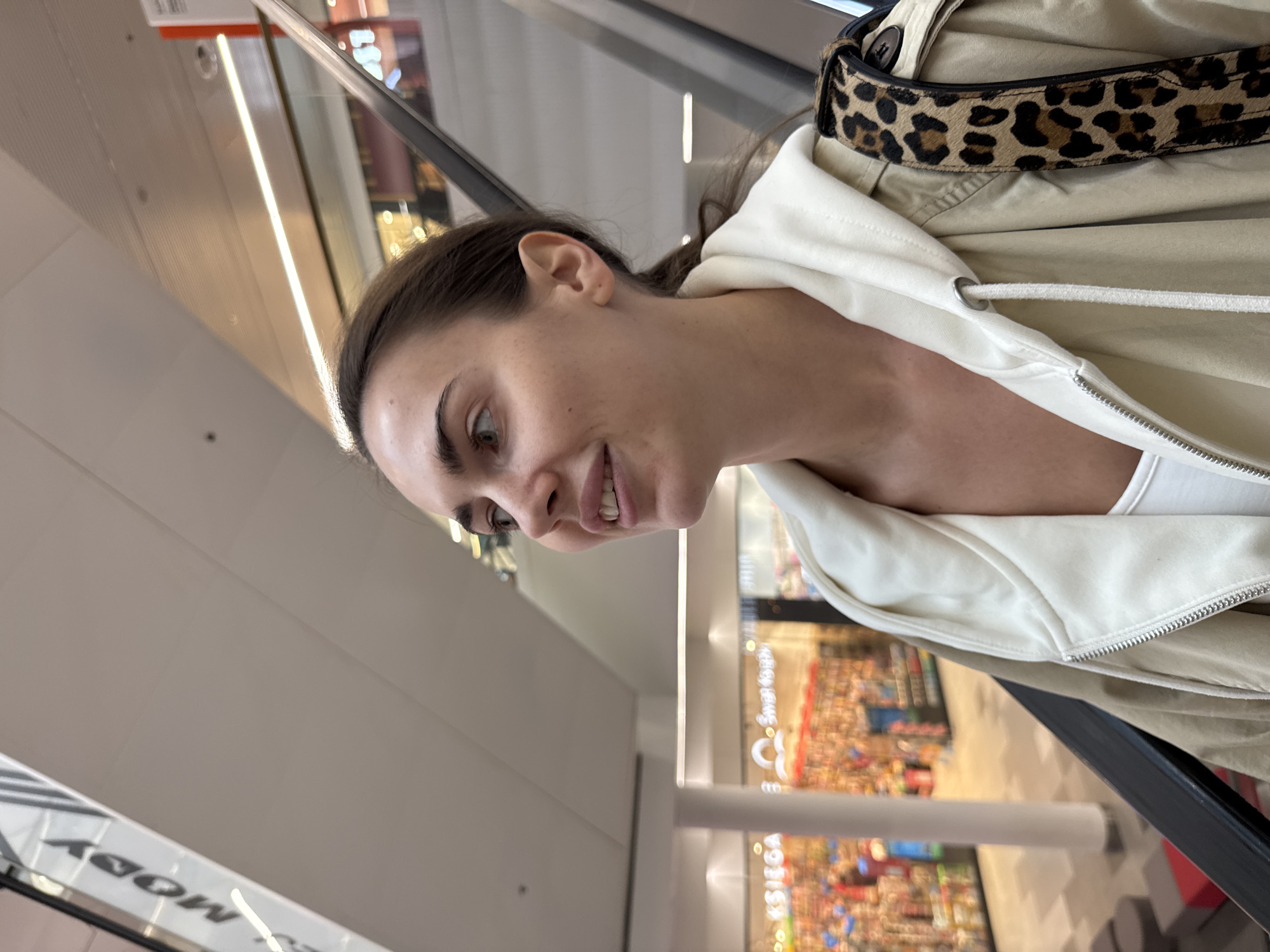 A young woman is captured in a close-up, portrait-oriented shot, rotated 90 degrees to the left, while riding an escalator in a brightly lit indoor commercial complex, likely a shopping mall in Warsaw, Poland. She is seen from the chest up, with her dark hair pulled back and distinct dark eyebrows framing her light-colored eyes. A gentle smile graces her face, revealing a glimpse of her teeth as she looks slightly downwards and to her left.
She is dressed in layers, including a light-colored (possibly white or off-white) hooded top or jacket with a visible drawstring, worn under a beige or khaki-colored outer jacket. A prominent leopard print strap, likely belonging to a bag, is visible over her right shoulder (relative to the image's orientation).
The background is a blurred but discernible interior of a multi-level building, showing the diagonal lines of escalators and architectural elements. Bright, linear light fixtures illuminate the ceiling. Below her, other levels of the mall are visible with various retail displays, suggesting numerous shops. Visible text on signs includes "MODY" (Fashion), seen in reflection, and "KSIEGA" (Book), suggesting a bookstore, further supporting the context of a shopping environment in Poland. The image captures a casual moment during what appears to be a shopping trip.