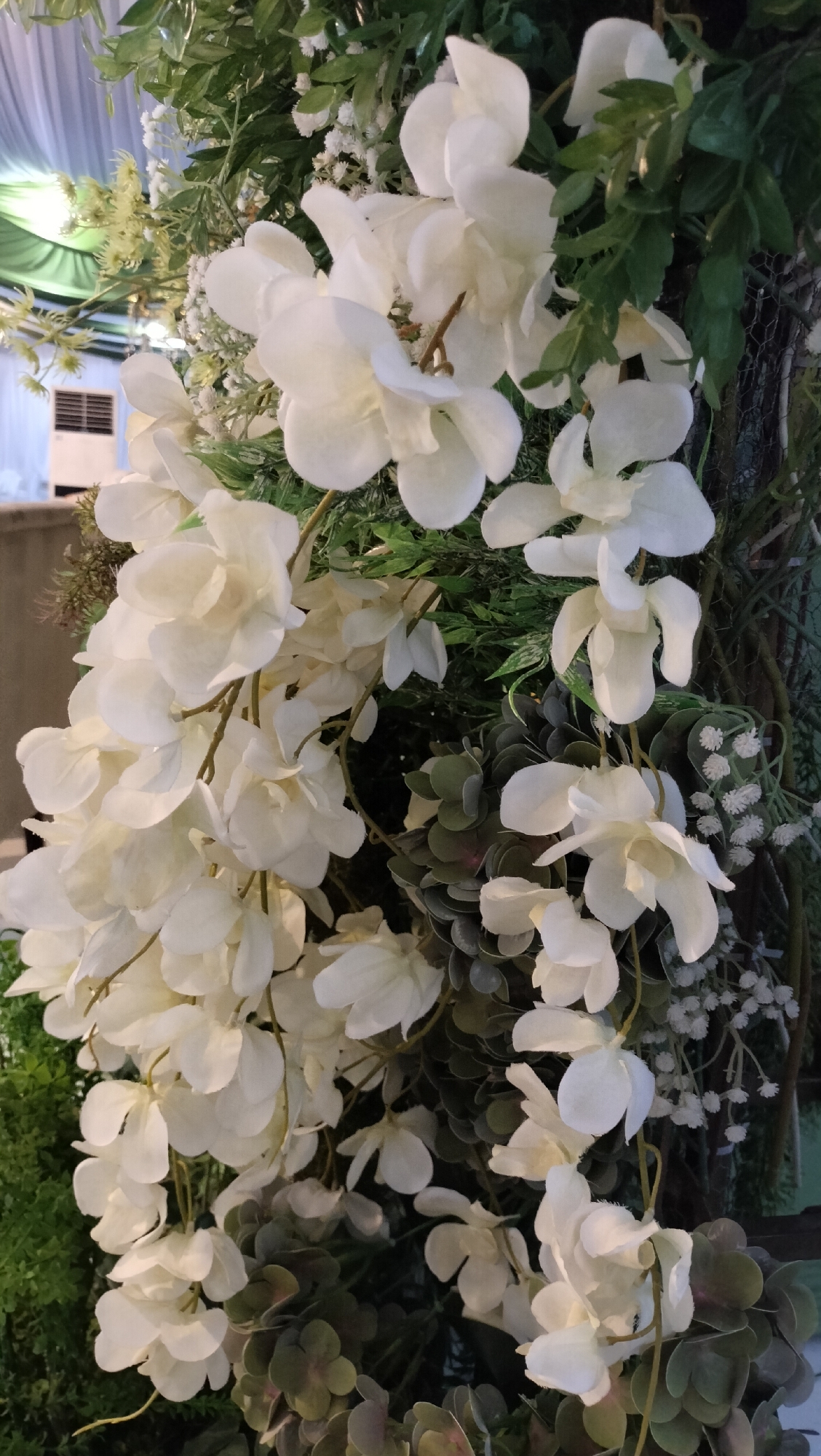 This is a close-up, eye-level shot of an artificial floral arrangement, likely a wedding decoration or event backdrop, featuring numerous white orchid-like flowers cascading downwards. The flowers are densely clustered and have delicate, layered petals. Interspersed among the white flowers are various green artificial leaves and foliage, some appearing as larger, broad leaves, while others are smaller and more delicate, creating a lush and voluminous display. In the background, out of focus, are glimpses of what appear to be event decor elements, including draped white fabric and possibly a white air conditioning unit. The lighting is soft, casting subtle shadows and highlighting the textures of the artificial petals and leaves. There is no visible evidence to suggest this specific scene is located in Lhokseumawe, Indonesia, or any specific city.
