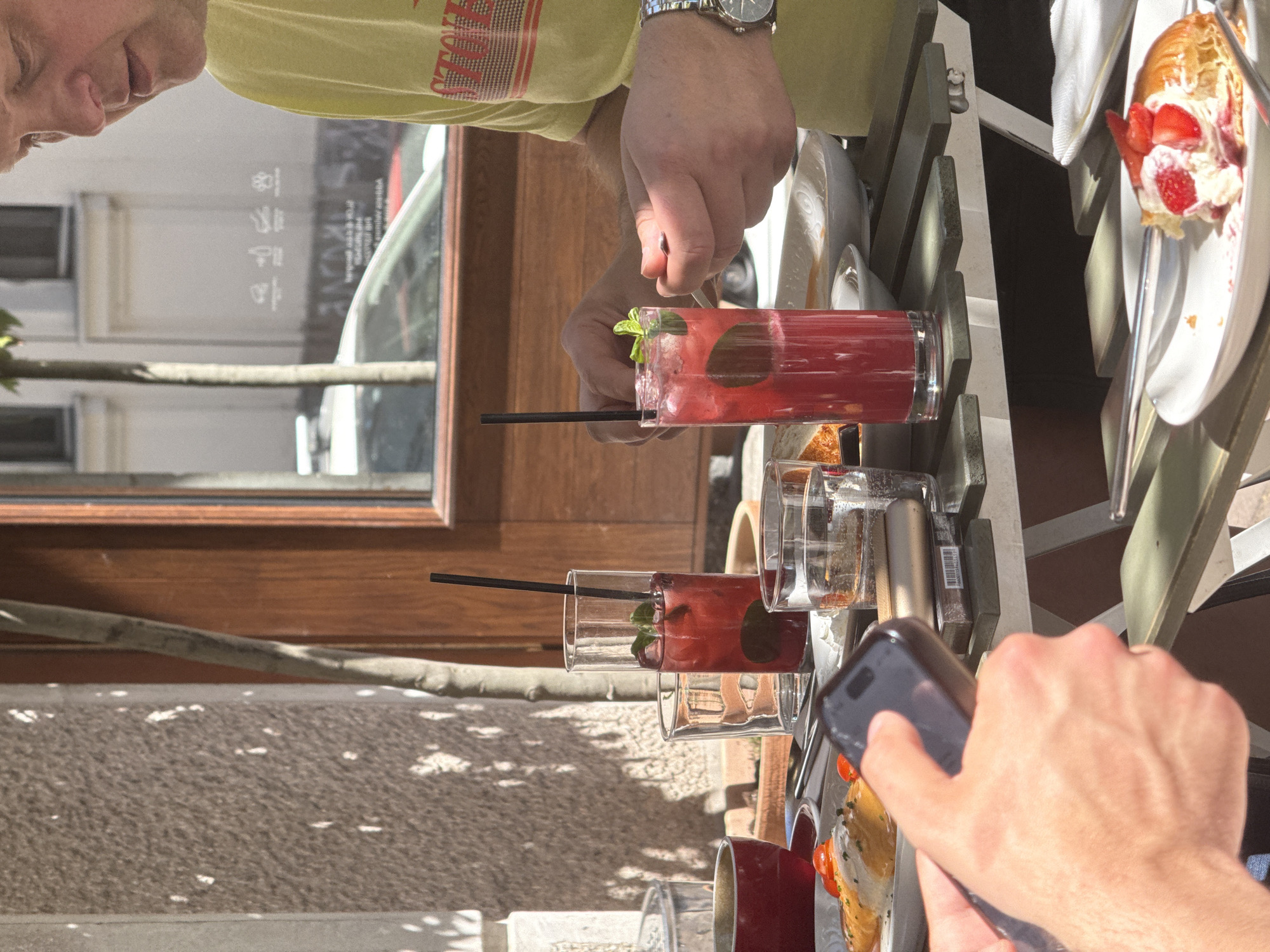 The image shows a restaurant scene in Belgrade, Serbia, focusing on a table with drinks and food. Two people are visible, one male wearing a yellow t-shirt with a watch on his wrist is handling a tall red beverage with a black straw and fresh mint garnish. The other person is taking a picture of the table with their cell phone. There are two glasses of the same red drink on the table, along with plates, cutlery, and a pastry topped with strawberries and cream. The setting is outdoors. A window or door is visible behind the table, reflecting the interior of a building, likely the restaurant itself. The table is gray. The ground is shaded and light-colored.