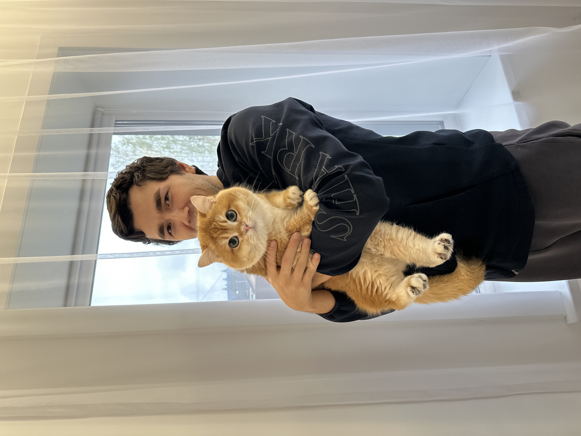 A man is holding an orange cat in his arms. They are standing in front of a window with a white curtain. The man is wearing a black hooded sweatshirt and gray pants. The cat has large eyes. The location is likely Молодёжный, Russia.