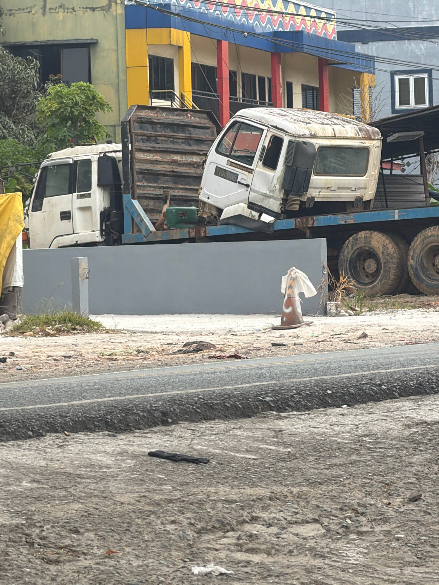 The image captures a scene on Jalan Ahmad Yani in Balikpapan, Indonesia, a commercial area with multi-story buildings. A flatbed truck is parked on the side of the road, carrying what appears to be the disassembled cabin of a white truck. The cabin is damaged and appears weathered, with rust visible on its surface. A portion of the original truck's chassis is also visible, loaded onto the flatbed. In the foreground, a traffic cone, partially covered by plastic sheeting, rests on the ground near a newly constructed grey wall. The road surface in front of the wall shows signs of wear and tear. The environment suggests a construction or dismantling site.