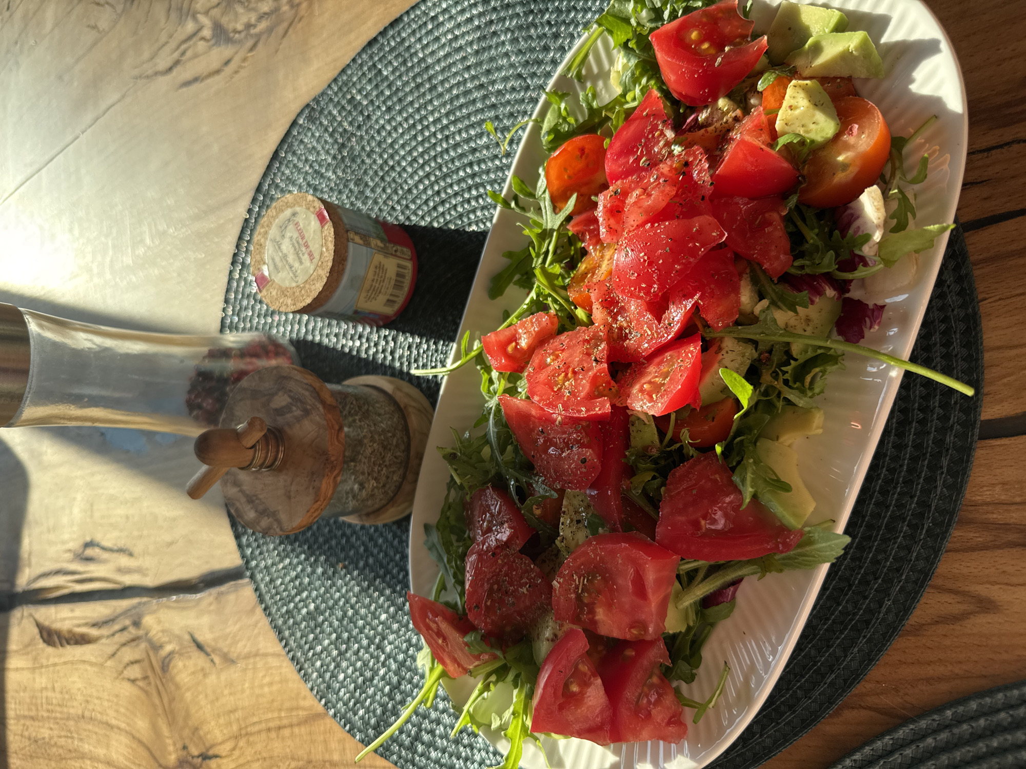 The image shows a white plate filled with a salad consisting of arugula, chopped tomatoes, and avocado. The salad is topped with spices. Beside the plate is a wooden pepper mill and a small jar with a cork lid. The items sit on a textured gray placemat, which rests on a wooden surface. The setting is indoors with bright, directional sunlight casting shadows. The location and any connection to Steklyannyi, Russia, are not discernible from the image.