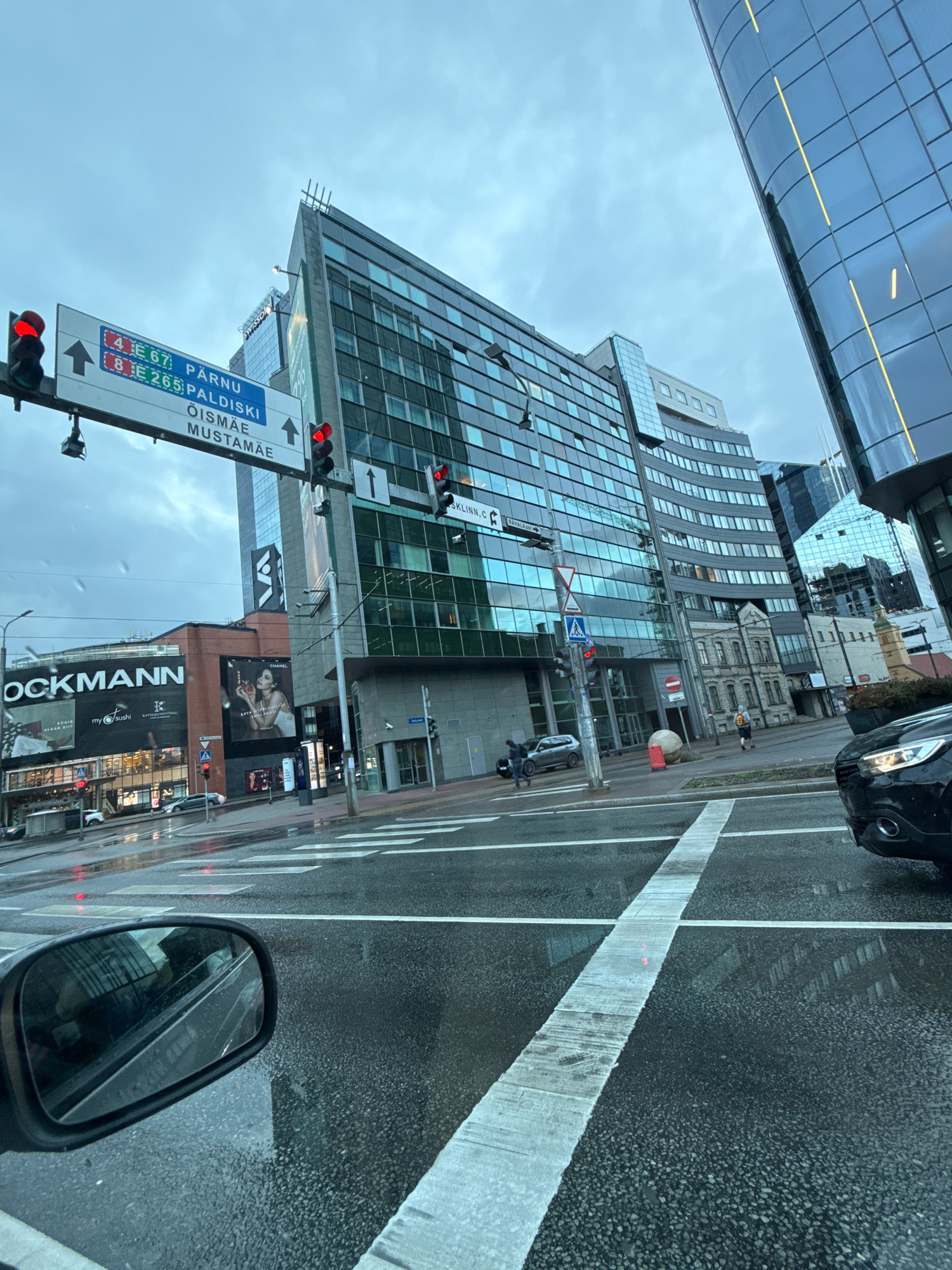 The image depicts a street scene in Tallinn, Estonia, on a cloudy, wet day. The perspective is from inside a vehicle, looking forward and slightly to the right. In the foreground, the wet asphalt road with white crosswalk markings is visible, along with the side mirror of the vehicle, reflecting the surrounding buildings.
Dominating the midground and background are modern glass-facade buildings, characteristic of a city center. One prominent building on the right has a curved glass exterior with vertical yellow lighting. To the left, a large building with a signage that reads "OCKMANN" is visible. Above the street, a traffic light is displaying red. A road sign is also present, indicating directions to "PÄRNU E 67", "PALDISKI E 265", and "ÕISMÄE MUSTAMÄE". Traffic lights are also visible on the signpost.
In the distance, several cars are on the road, and a few pedestrians can be observed. The overall environment suggests an urban setting with commercial and office buildings, and the wet conditions indicate recent or ongoing rain. The architectural style of the buildings, particularly the modern glass structures, places this scene within a contemporary business or commercial district. The specific street signs help to identify the location within Tallinn.