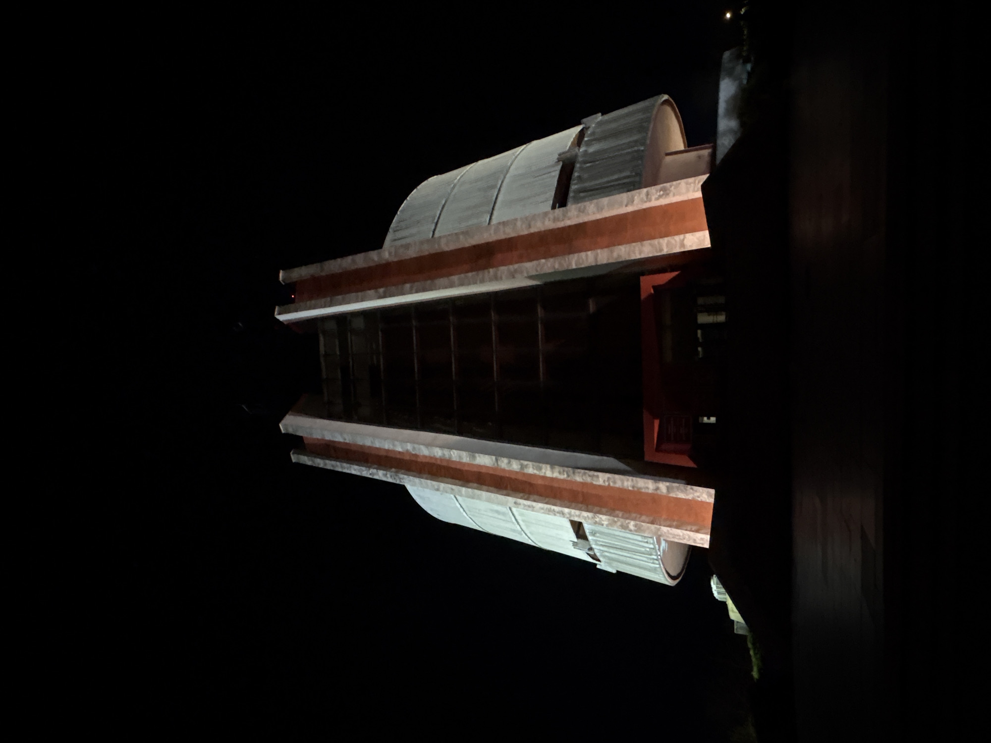 The image shows the Pleven Panorama in Pleven, Bulgaria at night. The building is constructed of gray panels supported by red beams. It consists of two parallel, horizontal structures. The lighting makes it appear reflective. The background is completely dark. There are no people in the image.