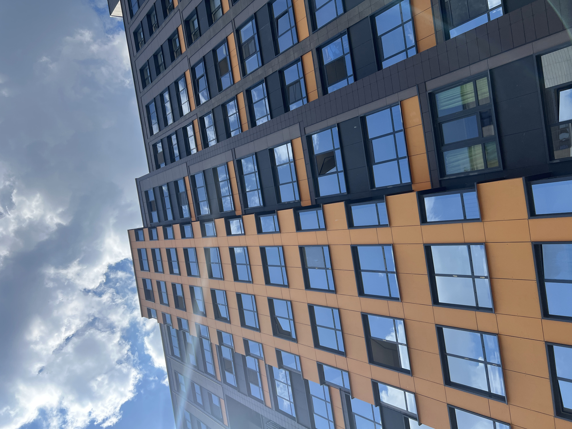 This image captures a modern high-rise building from a low-angle perspective, looking upwards towards the sky. The building is characterized by two distinct facade sections: one predominantly dark grey or black, and the other a vibrant orange or tan. Both sections are uniformly covered with numerous rectangular windows, each framed in black.
The windows prominently reflect the bright blue sky and scattered white, fluffy clouds, creating a dynamic interplay of light and color that merges the building with its surroundings. Some reflections are so clear that they almost appear as extensions of the sky itself, while others show subtle variations in light and shadow. A noticeable sun flare or glare is visible in the lower portion of the image and within some window reflections, indicating a sunny day.
The architectural style is contemporary, with clean lines and a multi-tiered or stepped design visible in the building's structure. The overall impression is one of modern urban development against a clear daytime sky. There are no people or specific activities depicted. The scene is set under clear weather conditions, and based on the provided context, the location is Saint Petersburg, Russia. No visible text is present in the image.