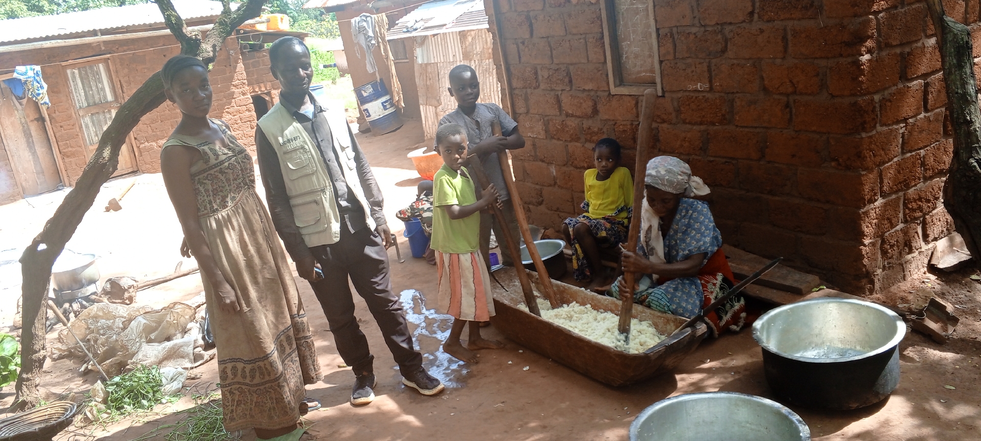 In Makungu, Democratic Republic of the Congo, a group of people stands outdoors. A young woman in a long dress stands to the left of a man wearing a button-down shirt and khaki vest. Three children stand near a woman who is working in a wooden container. One child is holding a long wooden stick. A woman wearing a headscarf and patterned clothing is pounding food inside the container with a pestle-like tool. Two metal pots are in the foreground. The surroundings are rural, with buildings constructed of brick and corrugated metal. There are trees and foliage. The ground is dirt.