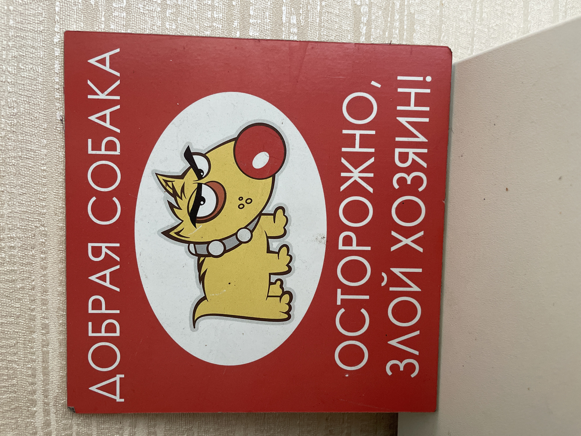 A square, red sign with white text and a cartoon dog is displayed on a textured, light-colored surface. The text at the top reads "ДОБРАЯ СОБАКА," which translates to "GOOD DOG." Below this, a stylized yellow dog with a large red nose, a studded collar, and an aggressive expression is depicted within a white circle. The bottom portion of the sign features the text "ОСТОРОЖНО, ЗЛОЙ ХОЗЯИН!", translating to "CAUTION, ANGRY OWNER!" The sign is positioned on a light-colored surface, possibly a table or shelf, against a backdrop of a textured wall. The lighting is even, and the image is captured from a slightly elevated angle. There is no information available that would allow for determining the location of this sign in Sochi, Russia.
