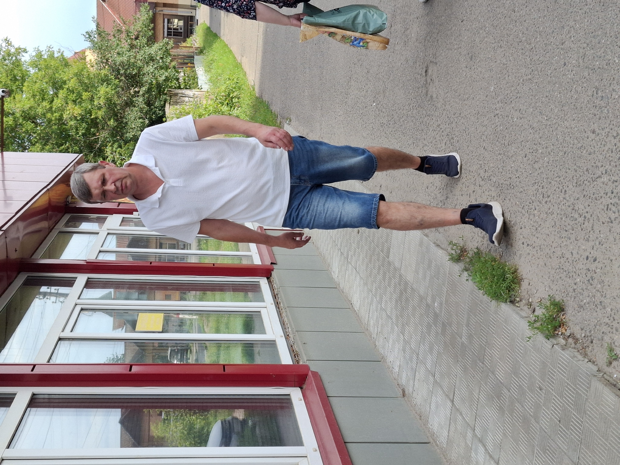 An adult male with short gray hair, wearing a white polo shirt and blue denim shorts, walks on a paved sidewalk. He is walking away from the camera towards the right side of the frame. To his left is the exterior wall of a building with large windows. Green grass and trees are visible in the background. A woman with a floral patterned shirt is walking in the same direction in front of him, carrying a bag and a flat wooden object. The weather appears to be daytime.