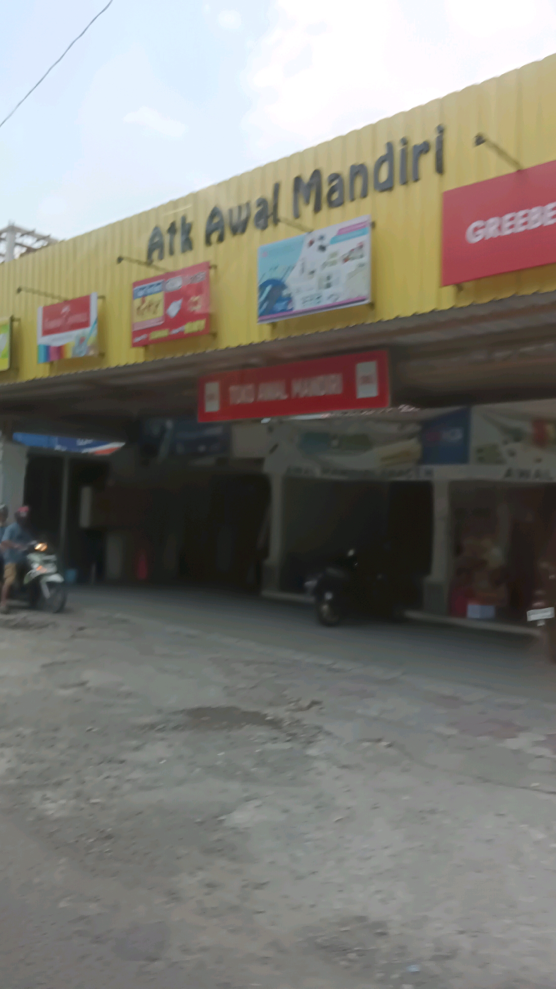 The image shows a storefront in the city of Sragen, Indonesia. The storefront has a yellow facade with a black sign that reads "Atk Awal Mandiri". There are several advertisements displayed on the facade, including one that reads "GREEBE" in red. Below the main sign, a red banner displays "TOKO AWAL MANDIRI" in white text. In front of the store, a person is riding a scooter. The ground in front of the store is paved with gravel and dirt. The sky is visible in the background. The overall scene suggests a commercial area with shops and businesses.