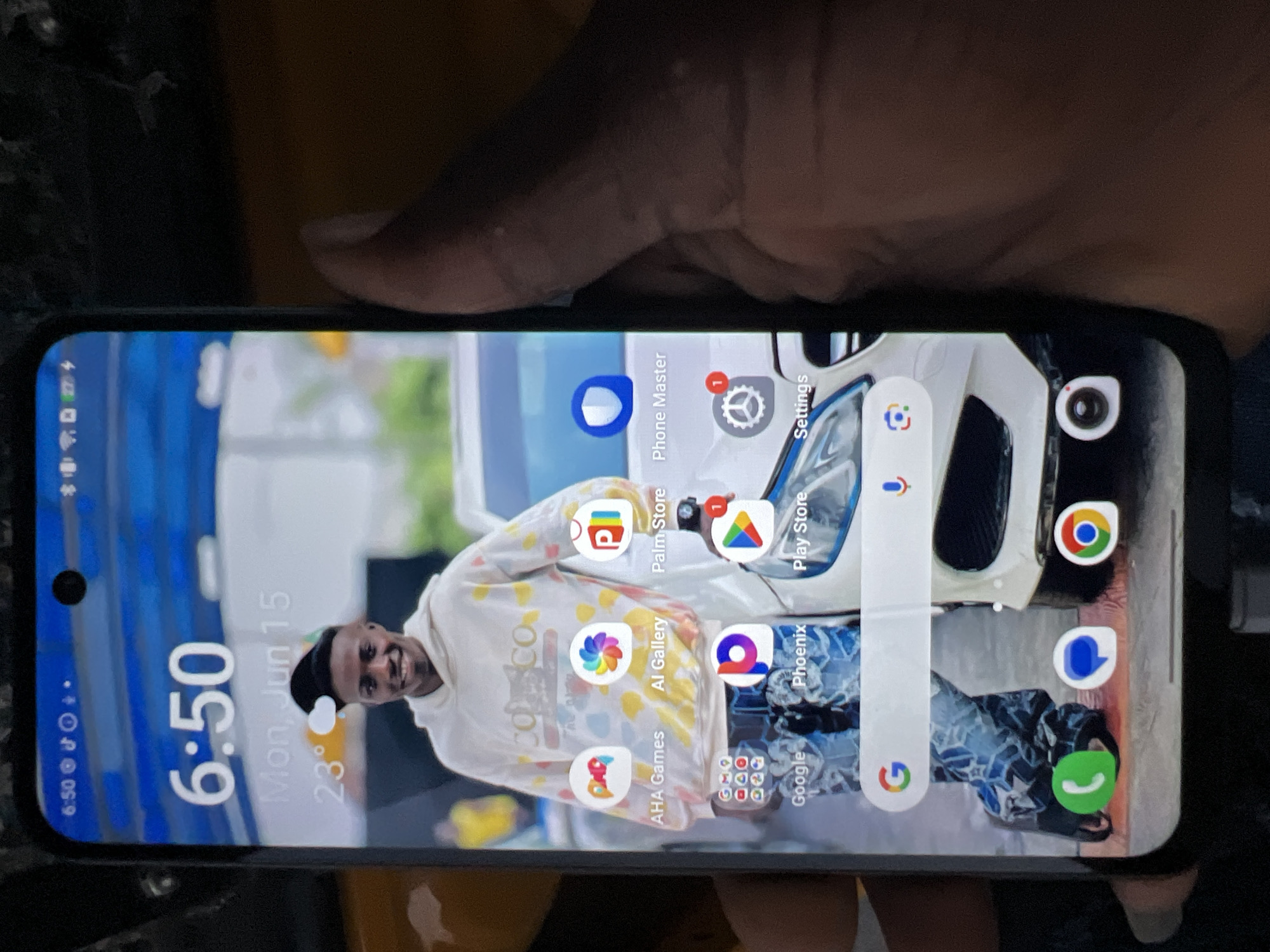 A black smartphone displays a lock screen with a time of "6:50" and the date "Mon, Jun 15" with a temperature of 23 degrees Celsius. The background features a young Black man wearing a white hooded sweatshirt with a colorful tie-dye pattern and a black beanie. He is smiling and looking at the camera. The lower portion of the screen shows various app icons including AHA Games, AI Gallery, Palm Store, Phone Master, Google, Phoenix, and Play Store. A Google search bar with a microphone icon is also visible. The phone is held by a person whose hand is partially in the frame on the right side of the image. The background appears to be outdoors with a car partially visible. The image quality is moderate. There is no specific information to indicate the location in the city of Orifite, Nigeria.