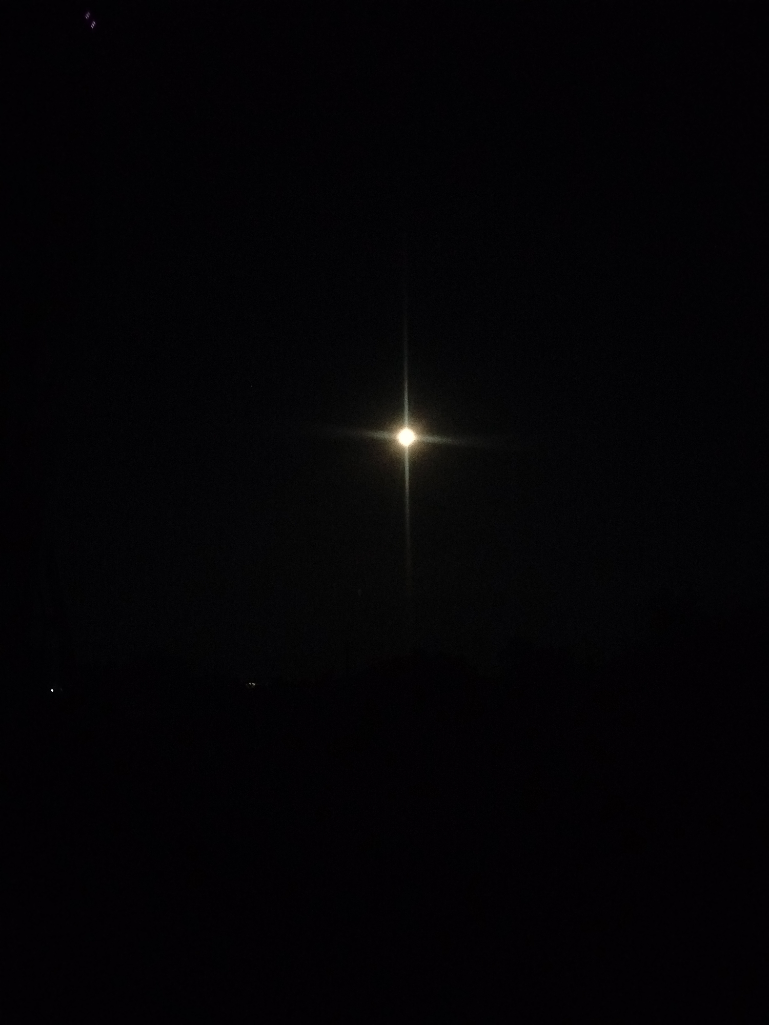 The image depicts a night scene, likely taken outdoors in Kostiantynivka, Ukraine. The dominant subject is a very bright, artificial light source in the center of the frame, casting a strong glare and a distinct starburst effect. This light is likely a street lamp or a similar overhead fixture. The background is almost entirely black, indicating nighttime. There are faint hints of distant lights or structures along the bottom horizon, suggesting a populated area, possibly a town or city.
In the upper left corner, there are a few small, indistinct purple or pinkish shapes, possibly reflections or artifacts. Two very small, faint dots of light are visible in the lower left quadrant. The overall impression is one of darkness with a single, powerful light source dominating the scene. There is no clear indication of weather conditions, but the darkness implies it is not daytime. The scene appears still and quiet, with no visible people or active events. The focus is on the stark contrast between the intense light and the surrounding darkness.