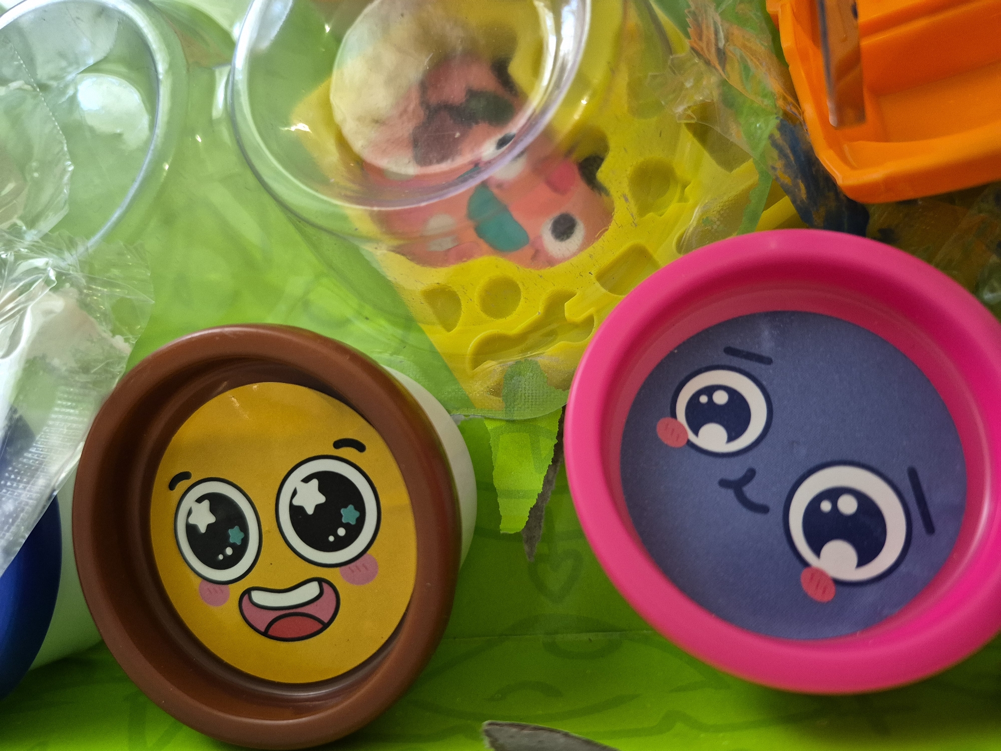 This close-up image showcases a collection of colorful children's play items set against a bright green background, likely a playmat or tablecloth, in an indoor setting in Yerevan, Armenia.
The primary subjects are two round containers, possibly for play-doh or craft supplies, featuring distinct cartoon faces. On the left, a container with a brown rim presents a cheerful, bright yellow face with large, sparkling eyes (depicting white stars and blue accents), rosy pink cheeks, and a wide, open-mouthed smile with a pink tongue. To its right, a container with a fuchsia pink rim displays a blue/purple face. This face has large, round eyes with white pupils, small red blush marks, and a subtle curved line for a mouth, conveying a curious or slightly shy expression.
In the upper portion of the frame, a clear plastic container is visible, housing a colorful, possibly sculpted item, like play-doh, which appears to be pink, orange, and blue/green. This clear container rests on a bright yellow, textured plastic surface that features a grid of circular indentations. Partially visible in the upper right corner is a piece of bright orange plastic, possibly part of a toy or tool, with visible ridges.
The overall scene is vibrant and suggests a playful atmosphere, characteristic of a child's activity space. There is no visible text in the image.