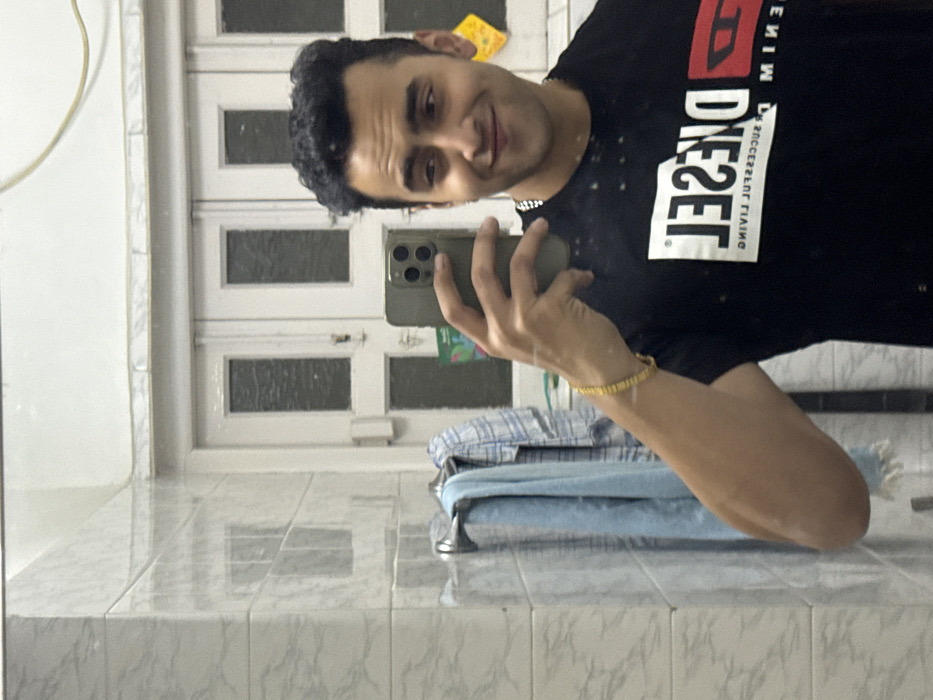 A person wearing a black Diesel t-shirt is taking a selfie in a bathroom. The person is holding a phone with three camera lenses. The bathroom has white tiled walls and a white door with square windows. A stack of folded blue and white clothing is visible on a surface behind the person. A gold bracelet is on the person's wrist. A small yellow object is stuck to the door frame behind the person.