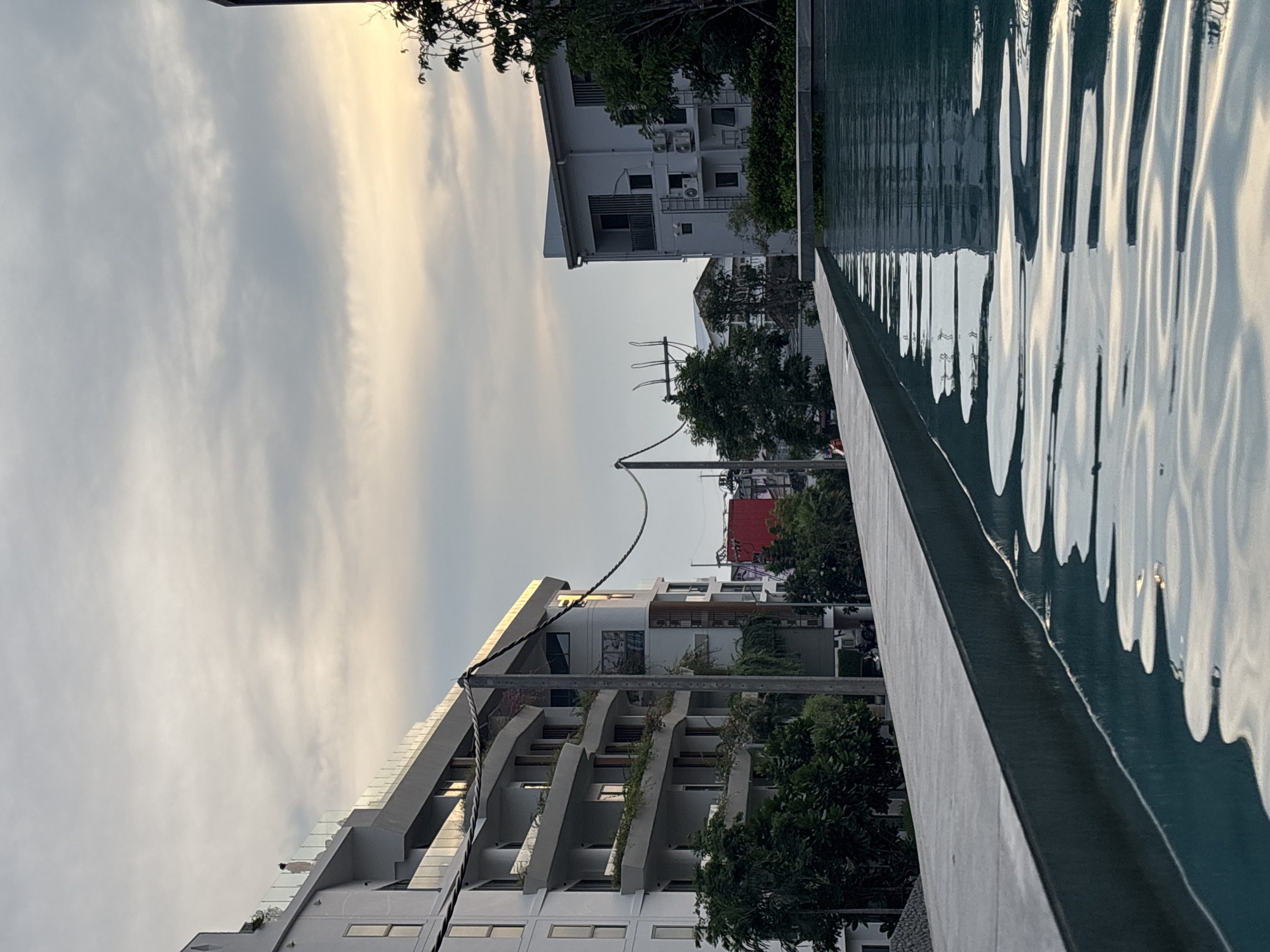 This photograph presents an outdoor scene in Choeng Thale, Thailand, featuring a swimming pool alongside various buildings and a sky suggesting either sunrise or sunset. The image is oriented vertically but depicts a horizontal landscape.
In the right foreground, a swimming pool with dark blue-green water displays prominent ripples, reflecting the sky and nearby structures in distorted, wavy patterns. A light grey concrete edge borders the pool.
On the left side of the frame, several buildings are visible. One prominent, modern multi-story building features balconies with glass railings and some integrated greenery. Adjacent to it, another building appears simpler, with white walls and exposed air conditioning units. Lush, green tropical foliage and trees are interspersed among the buildings, adding a natural element to the urban environment. A utility pole with multiple overhead wires cuts across the mid-ground, a common sight in the region.
The background is dominated by a partly cloudy sky. On the left, the sky is a soft, muted grey, transitioning towards a warm, golden-yellow hue on the right. This warm glow, combined with the soft lighting, suggests the time of day is either early morning or late afternoon.
There are no people visible, and no specific activity is taking place, contributing to a calm and serene atmosphere. The architectural styles, the utility pole, and the dense tropical vegetation serve as strong location cues for Southeast Asia, aligning with the given context of Choeng Thale, Thailand. No visible text is present in the image.