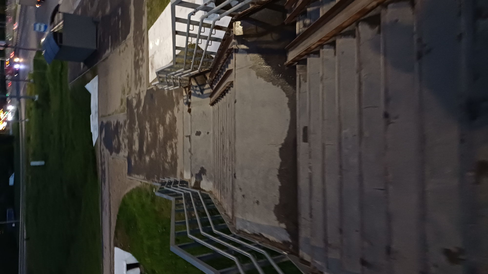 The image shows a concrete structure with a staircase and metal railings. The location appears to be in an urban area, likely in Obukhovo, Russia, based on the information provided. To the left of the structure, there is a grassy area with trees and streetlights visible in the distance, suggesting a street or park. The photo seems to have been taken during the evening or night, as indicated by the streetlights and darker lighting. The concrete appears to be damp or wet, indicating rain or recent cleaning. The scene is devoid of people.