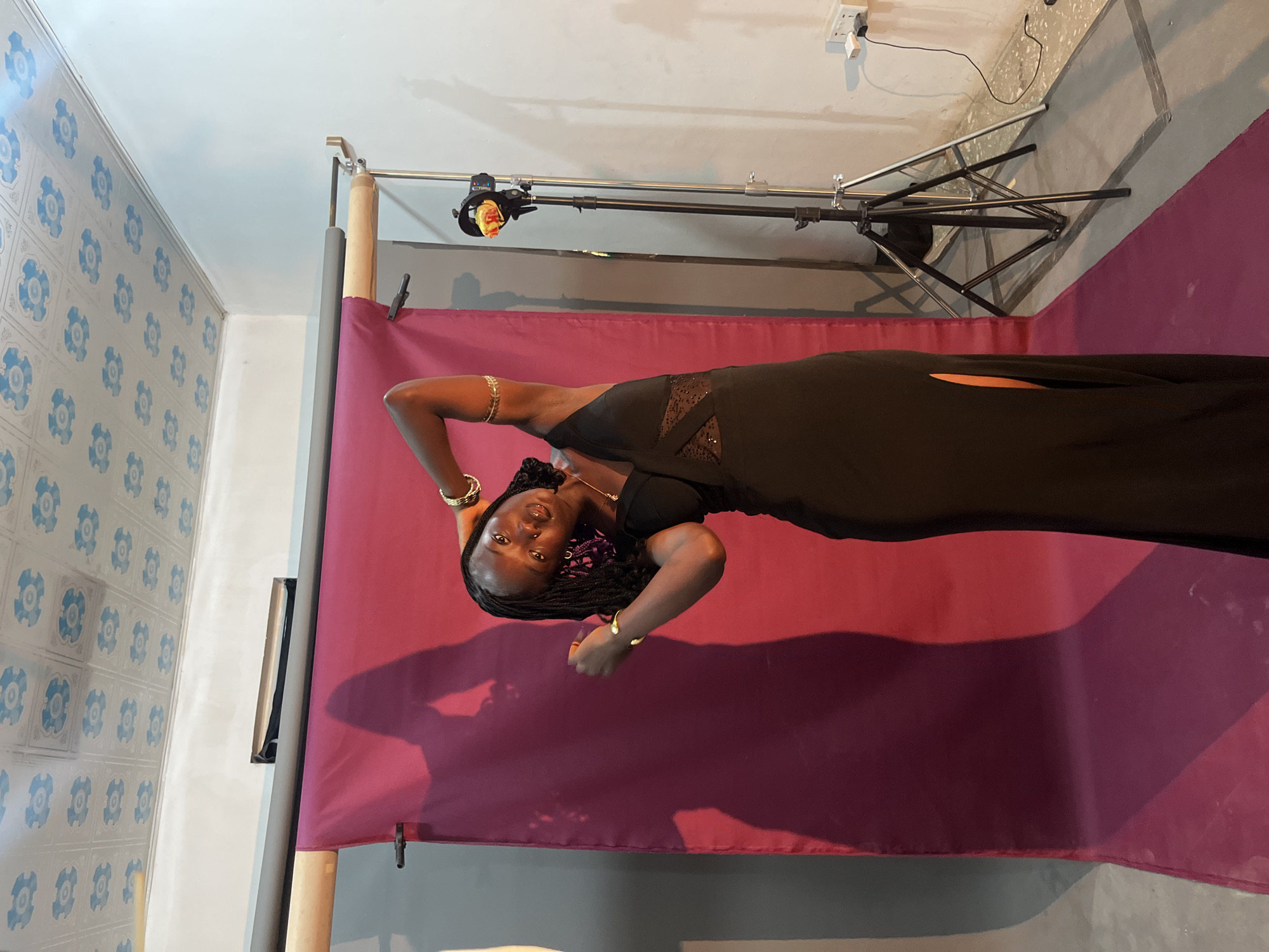 An outdoor photo shoot is taking place in what appears to be a makeshift studio located on a rooftop in Gura Top, Nigeria. The backdrop is a deep magenta fabric, tautly stretched between two vertical poles. To the right of the backdrop, a portable studio light on a tripod casts a dramatic shadow of the subject onto the fabric. The subject, a young Black woman with braided hair, is wearing a long, elegant black dress with a thigh-high slit and an asymmetrical neckline featuring a sheer, patterned insert. She is posing with her arms behind her head and her body tilted, looking directly at the camera with a confident expression. She is adorned with gold bracelets on both wrists and a delicate gold necklace. The ceiling above is paneled with a decorative pattern of blue and white floral motifs, suggesting an indoor or covered rooftop setting. The floor is concrete, marked with faint gray lines. A black cable runs from a wall socket to the tripod. The overall atmosphere is one of a professional or semi-professional photo session.