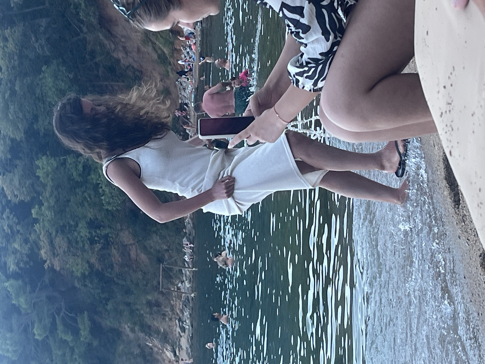 In Budva, Montenegro, on a pebbly beach leading into the Adriatic Sea, a young woman with long, dark, curly hair is standing at the edge of the water. She is wearing a white tank dress and is lifting the hem of the dress with her right hand. Her left hand is holding a dark-colored cell phone, which she is using to take a photo of herself or the scene in front of her. To her right, another young woman is seated, facing away from the camera, and is also holding a cell phone, seemingly taking a picture. In the background, the sea is populated with numerous people swimming and wading, and a densely wooded hillside rises behind the beach, dotted with rocky outcrops. The water is a deep green, with light reflecting off its surface.