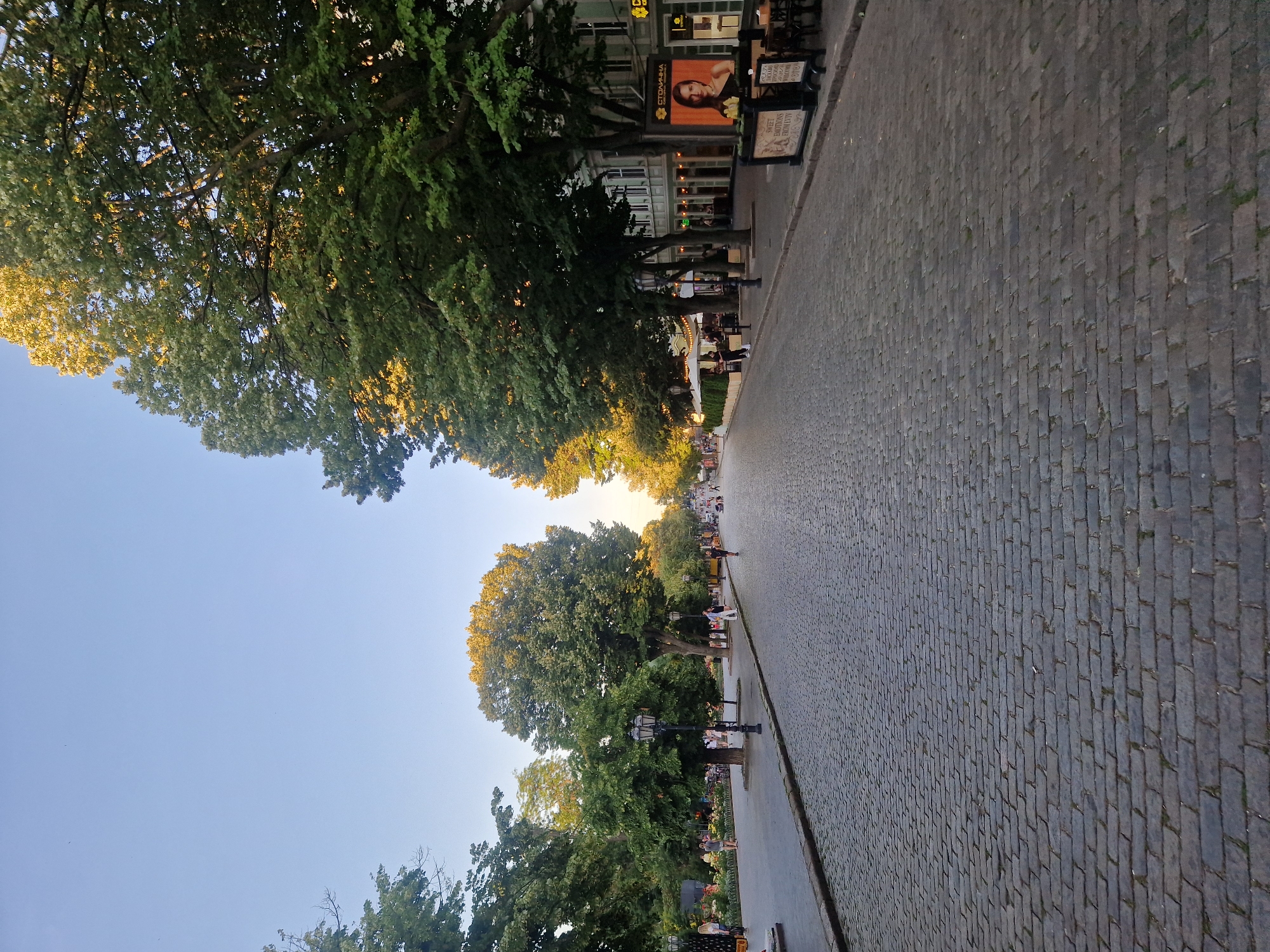 This is a vertical, high-angle shot of a cobblestone street lined with trees on one side and buildings on the other. The sun appears to be setting, casting a warm glow on the leaves of the trees and the sky. People are visible walking along the street and sitting at outdoor cafes. The scene evokes a peaceful, European atmosphere.
**Subjects:**
* **People:** Several individuals are walking on the sidewalk and street, some appear to be seated at outdoor cafes.
* **Objects:** Trees, lampposts, outdoor cafe tables and chairs, signs, cobblestones.
* **Scene:** A picturesque street scene, likely in a city or town.
* **Setting:** An outdoor urban environment with a prominent cobblestone walkway, mature trees, and buildings.
**Activity or Event:**
The image captures a relaxed, everyday scene of people enjoying an evening stroll and outdoor dining.
**Notable Details:**
* **Time of Day:** Late afternoon or early evening, indicated by the warm, golden light on the trees and sky.
* **Weather:** Appears to be clear and pleasant, with a bright blue sky.
* **Location Cues:** The cobblestone street and architecture suggest a European setting.
* **Emotions:** The overall mood is calm and serene.
**Visible Text:**
* A large advertisement board displays an image of a woman and text in what appears to be Cyrillic script, "СТОЛИЧНАЯ" (Stolichnaya).
* Another sign reads "Sweet Memories" in English, with additional text below it.