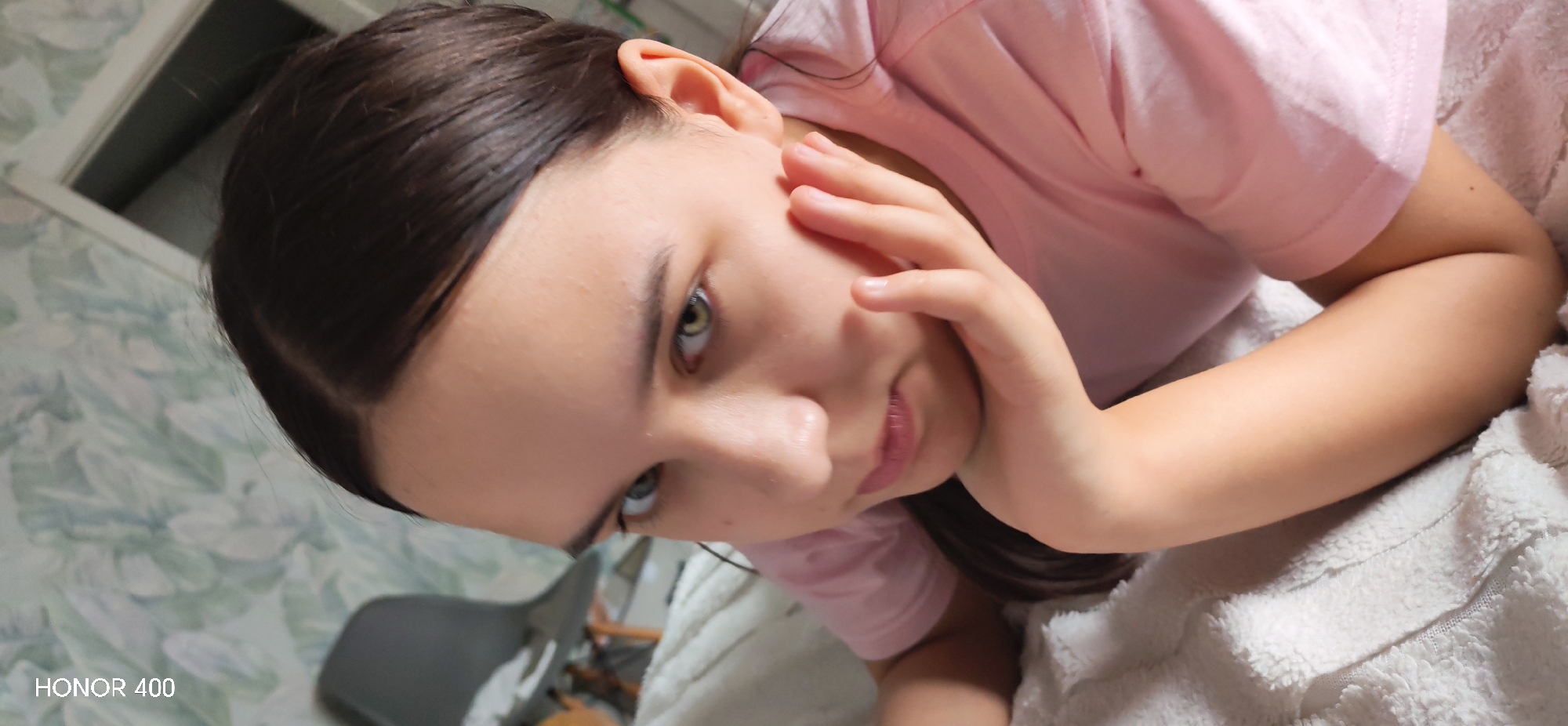 The media file presents a close-up portrait of a young person, oriented sideways, taken indoors. The subject, who appears to be female, has dark brown hair and light green or hazel eyes, gazing directly at the camera. She is wearing a light pink short-sleeved t-shirt. Her right hand is positioned gently against her cheek and jawline, suggesting a relaxed or contemplative pose.
The background reveals patterned wallpaper with light green and white leaf motifs. A dark grey chair is partially visible on the left side, and a white frame, possibly a door or window, can be seen above it. The person appears to be resting on or covered by a white, fluffy blanket or towel. The lighting is consistent with an indoor setting. The bottom left corner of the image displays the text "HONOR 400," likely a camera watermark. The image does not contain specific visual cues to corroborate the provided location context of Noviy, Russia, depicting a generic indoor environment.