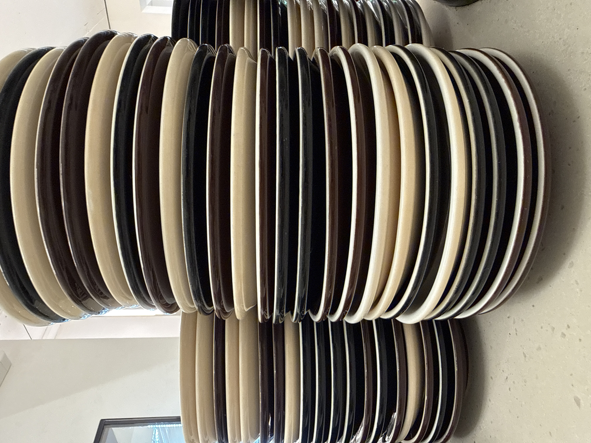 The image depicts multiple stacks of ceramic plates arranged on a light-colored, speckled countertop, likely within an interior space in Uzhhorod, Ukraine. The plates are primarily in three distinct colors: dark brown, beige/cream, and black/dark grey, stacked in an alternating pattern.
The most prominent stack, positioned right of center, is the tallest, featuring numerous plates arranged vertically. To its left, a shorter stack is visible, topped with beige/cream plates. A third stack, partially cropped on the far right, also follows the same color scheme. The plates exhibit a subtle sheen, indicating a glazed finish.
The countertop beneath the stacks is light grey or beige with dark speckles, consistent with a granite or quartz surface. The background consists of a plain white wall, with a dark-framed object, possibly a window or a framed picture, partially visible on the far left. The overall lighting is bright and even, suggesting an indoor environment such as a kitchen, dining area, or storage facility. No individuals are present in the frame.