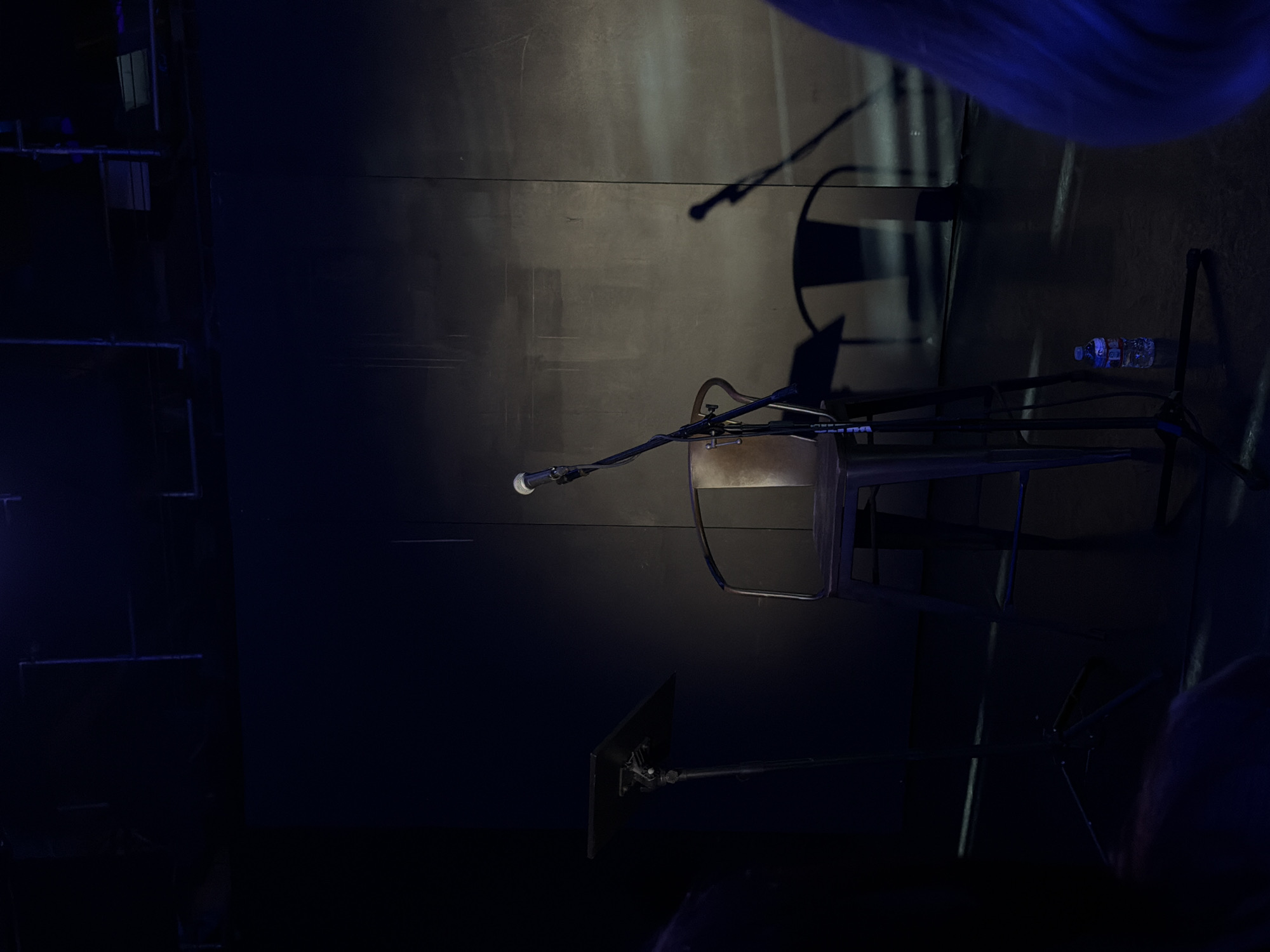 The image depicts a dimly lit stage, possibly in a small venue or studio, in Los Angeles, California, United States of America. The scene is set with a single stool positioned center-stage, equipped with a microphone on a stand. To the left of the stool, a music stand is visible, suggesting a performance or practice setting. The background is a dark, textured wall, with some ambient lighting creating subtle shadows. A partially visible shadow of a microphone and stand is cast on the wall to the right of the stool. A plastic water bottle rests on the floor near the stool. The overall atmosphere is subdued and ready for a performance.