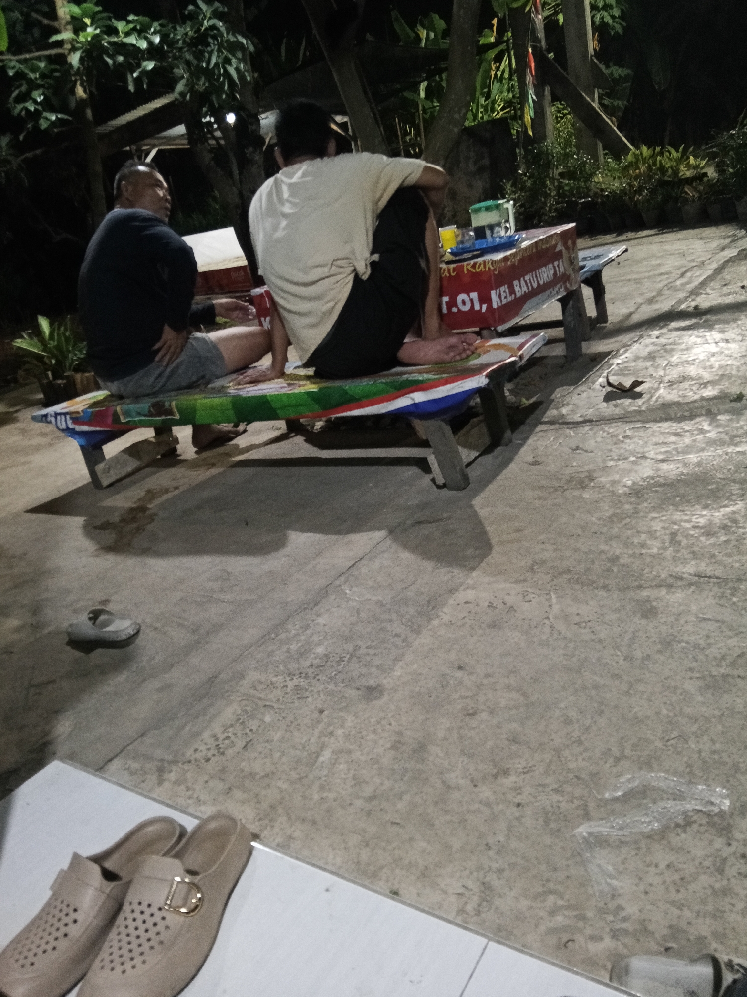 In Lubuklinggau, Indonesia, two individuals are seated on a long, narrow, decorated bench on a concrete surface. The person on the left is wearing a black long-sleeved shirt and grey shorts, with their left arm resting on their lap. The person on the right is wearing a white t-shirt and black shorts, with their back to the camera and their legs crossed. In front of them is a table with various items, including drinks in plastic cups and containers. The environment appears to be outdoors, with trees and plants visible in the background. The lighting suggests it is nighttime or dusk. Two pairs of shoes are visible in the foreground, one pair of light-colored slip-on shoes with a buckle and a single grey sandal on the concrete. A piece of clear plastic is also on the ground.