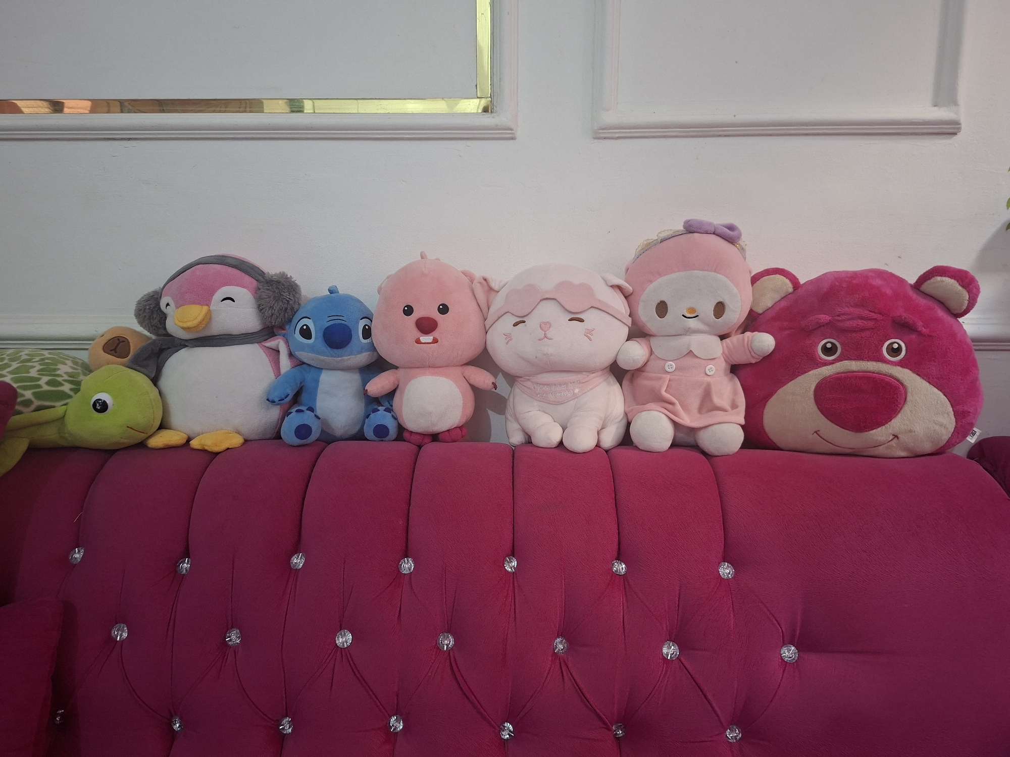 Six plush toys are neatly arranged in a row on the backrest of a vibrant magenta sofa. The sofa features deep tufting with clear, decorative buttons. From left to right, the toys include a green turtle, a white penguin wearing a pink and grey winter hat and scarf, a blue Stitch character, a pink pig-like character with a red nose, a light pink cat-like character with an eye mask and a bib (on which partial text like "YOU NEED ME" is visible), a light pink My Melody in a dress, and a large, dark pink Lotso bear from Toy Story. Most of the plush toys display smiling or peaceful expressions.
The sofa is placed against a bright white wall adorned with decorative molding, including a horizontal golden strip near the top and two rectangular golden-framed sections further up. The scene is brightly lit, indicating daytime indoors. The overall impression is one of coziness and playfulness within an indoor setting, likely a home in Ciseeng, Indonesia. No specific activity is taking place; the toys are arranged for display.