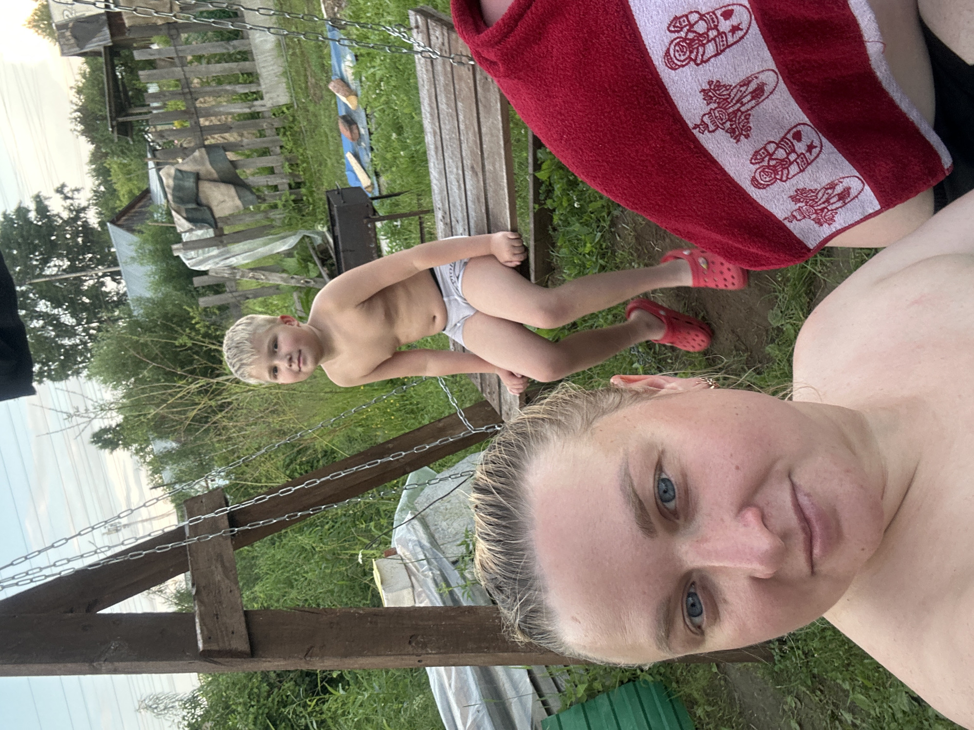This media file captures a candid moment of two individuals outdoors, possibly in a backyard or recreational area in Chaikovsky, Russia. The image is oriented vertically, with the top of the frame appearing on the left.
The primary subjects are two people: a young boy and an adult. The boy, in the foreground and slightly to the left, is wearing light-colored briefs and appears to be lying or sitting on a wooden structure, possibly a bench or part of a swing set, which is suspended by chains. He has fair hair and is looking towards the camera. The adult, occupying the right side of the frame, is a woman with light hair and blue eyes, also looking towards the camera. She is topless, with a red towel adorned with a white stripe featuring cartoonish figures draped over her shoulder. Red Crocs are visible on both of their feet.
The setting is a grassy, outdoor environment, with greenery and trees in the background. A wooden fence and some rustic structures, including what appears to be a grill and some tarpaulin or sheeting, are also visible, suggesting a casual, perhaps rural or suburban, location. The lighting suggests it is daytime, likely late afternoon or early evening, given the warm tones in the sky and the overall lighting. There are no discernible emotions or specific activities beyond posing for the photo. The scene appears relaxed and informal. No visible text is present within the image.