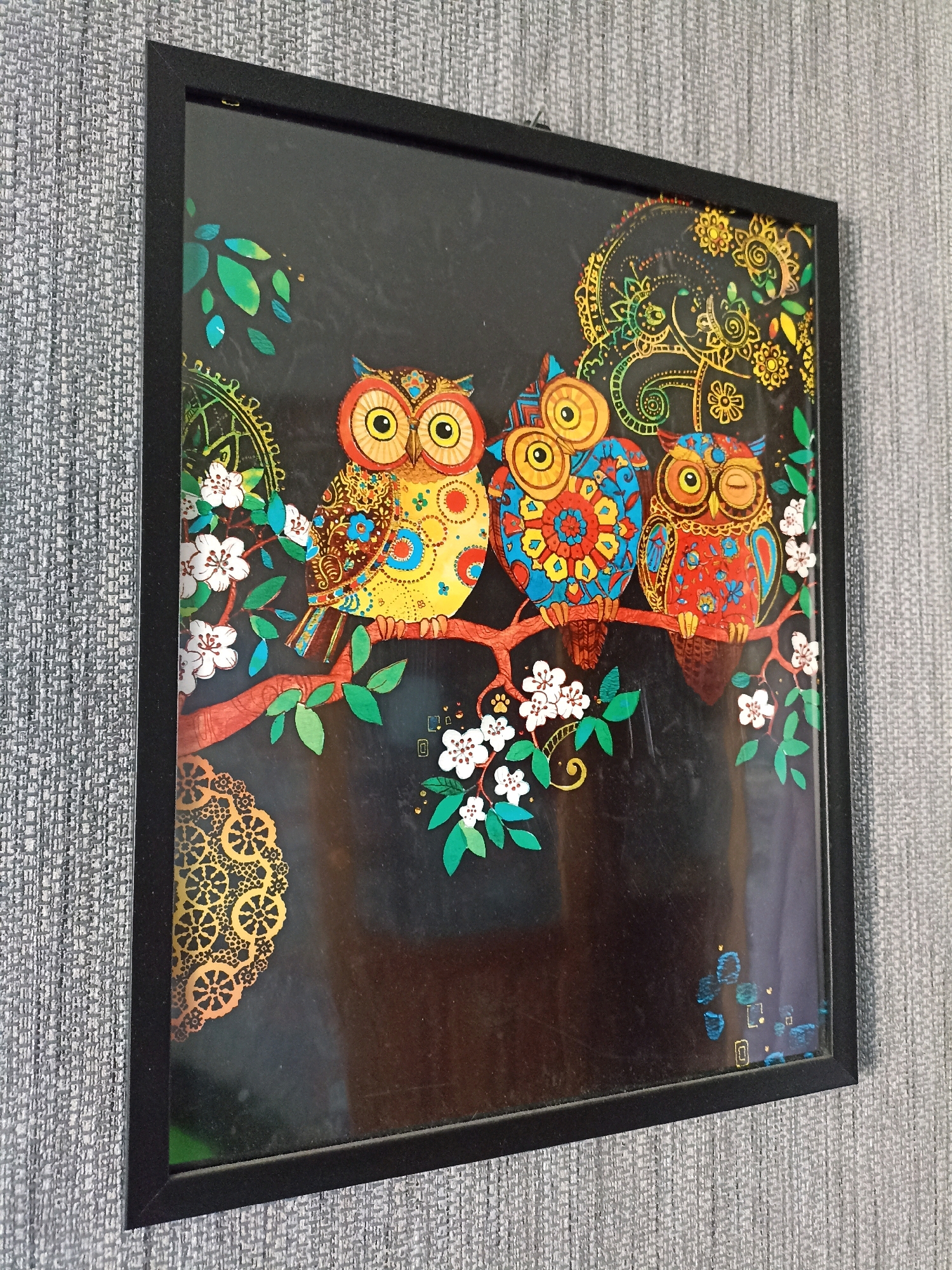The image shows a framed artwork featuring three vibrantly colored owls perched on a branch against a dark background. The owls are depicted in a whimsical, folk-art style with intricate patterns and bright hues of yellow, blue, and red. The branch is adorned with white blossoms and green leaves. Decorative golden motifs, resembling mandalas and swirling patterns, are also present in the background and on the owls themselves. The artwork is framed in black and hangs on a grey, textured wall. There are no people present in the image. The location in Novosibirsk, Russia, cannot be determined from the visual information provided.
