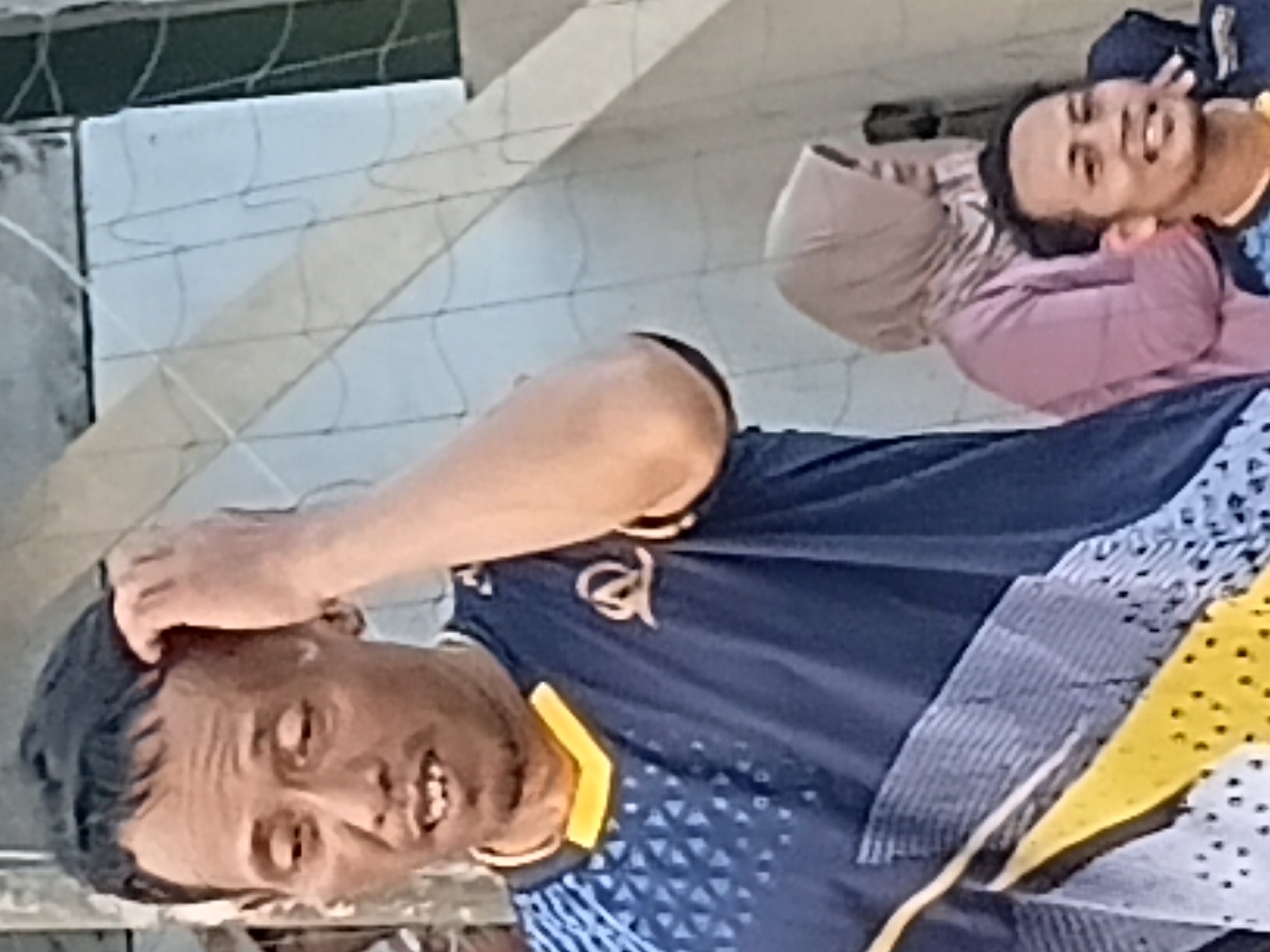 The image captures a medium shot of three individuals in what appears to be an outdoor setting, possibly a public space or sports venue, within the city of Margo Rukun, Indonesia. The primary subject, a man in the foreground, is wearing a dark blue athletic shirt with yellow trim and a patterned design on the left side. He has dark hair and is seen scratching his head with his right hand, with his mouth slightly open, revealing his teeth. To his right, and slightly behind him, is another man, also wearing a dark blue athletic shirt with yellow trim and a visible logo. This man is smiling and looking to his right. In the background, a woman is visible wearing a light pink headscarf and a pink top. She is looking to her left. The environment includes a net-like structure and a light-colored wall or building. The lighting suggests it is daytime.